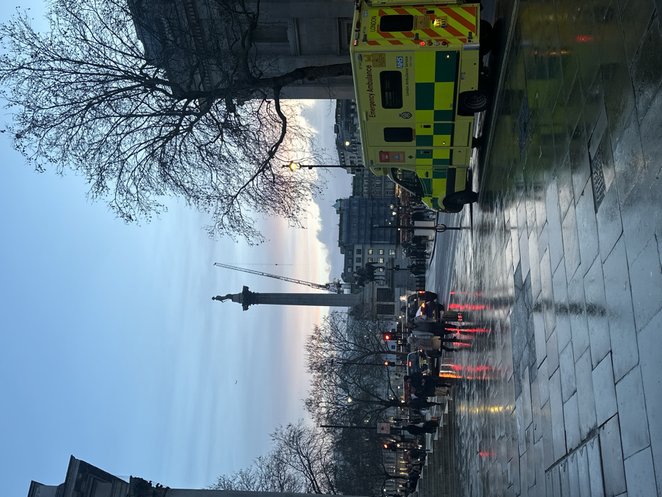 This media file captures a bustling street scene in London's Trafalgar Square, depicting an urban environment likely at dusk or dawn. The sky displays soft gradients of blue, transitioning to pale orange and pink hues near the horizon, indicative of the early morning or late evening light. The ground, paved with flagstones, is visibly wet and highly reflective, mirroring the surrounding lights and architectural elements.
In the foreground to the left, a large deciduous tree with bare branches partially frames the upper left corner of the scene. To the right, a bright yellow and green London Ambulance Service vehicle is prominently featured, facing away from the viewer. Visible text on the ambulance includes "Emergency Ambulance," "NHS," and "London Ambulance Service," along with the word "LONDON."
In the mid-ground, the iconic Nelson's Column, topped with its statue, stands tall as a central landmark. Beyond it, a large construction crane is silhouetted against the sky. The street is active with vehicles, including cars whose red taillights cast long, vibrant reflections on the wet pavement. Pedestrians are visible along the sidewalks and at what appears to be a crosswalk, adding to the sense of urban activity. Buildings with classical architectural styles line the background, contributing to the distinct London setting. The wet conditions and artificial lights reflecting create a dynamic and atmospheric view of the city.