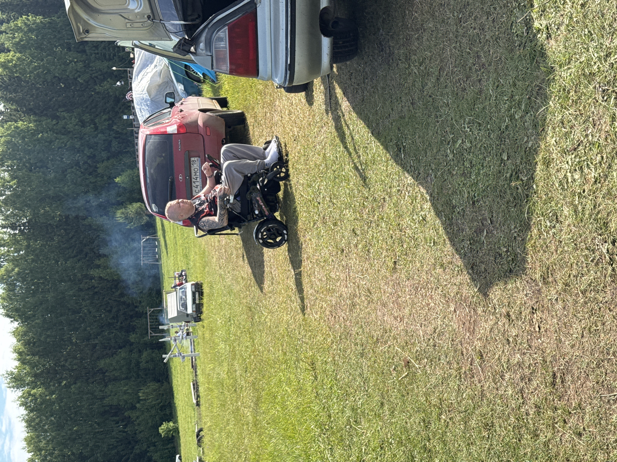 The scene depicts an outdoor gathering in a grassy field in Большая Кивара, Russia, under clear daylight conditions. In the mid-foreground, a light-skinned male with a shaved head and tattoos on his left arm sits in an electric wheelchair. He is wearing a dark t-shirt with red patterns, light grey sweatpants, and white socks or shoes, holding a control device for the wheelchair. He is positioned facing towards the left.
Behind the man, a red Lada Kalina hatchback is parked, displaying Russian license plate `C 414MO 18`. To the right of the man, the rear of a silver sedan with an open trunk is partially visible. Further into the midground, to the left of the red car, a white pickup truck is parked near a metallic structure of white poles. Behind these vehicles, a large white tarp-covered object is present. Smoke is observed rising from the left side of the midground, indicating a burning source.
The ground is covered in green and dry grass, with a well-trodden dirt path running diagonally from the bottom left corner towards the center. Long shadows are cast from the right side of the image towards the left. In the background, a dense line of tall green deciduous trees forms the horizon. Partially visible within the trees are two wooden swing sets, one towards the left and another towards the right. The sky above is light blue with sparse white clouds.