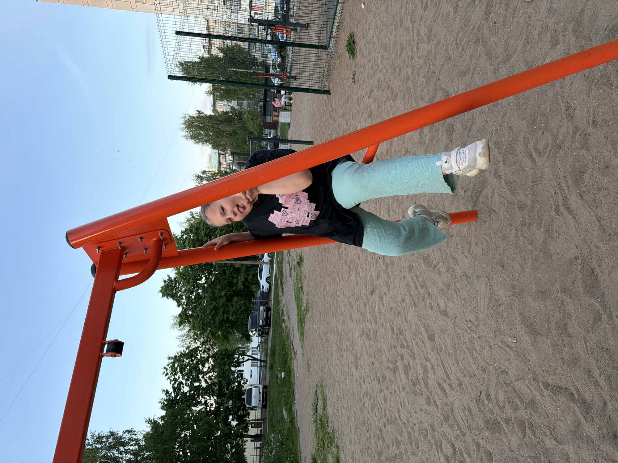 A child in a black t-shirt with pink decorations and turquoise pants hangs upside down on a bright orange outdoor exercise structure in a playground covered in sand. The child is smiling and sticking their tongue out. In the background are trees, grass, parked cars, and a fenced area. In the distance, a building can be seen. The location is Chaikovsky, Russia.