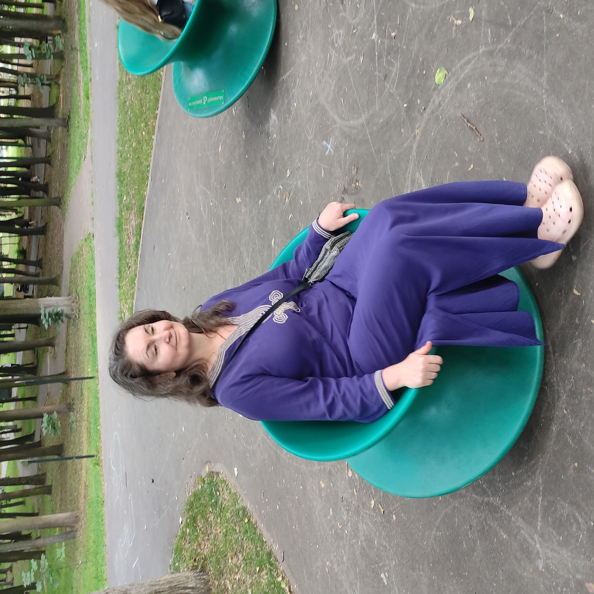 A medium shot captures a woman sitting in a green, curved, outdoor seating unit within a park. The woman, with medium-length brown hair, is wearing a long, dark purple dress with decorative trim around the neckline and cuffs. She is looking to her left with a slight smile. A small, dark-colored crossbody bag with a textured strap is slung over her right shoulder. Her feet are clad in light pink clogs. The seating unit is positioned on a paved area marked with white chalk drawings. In the background, trees line a path, and glimpses of other park-goers are visible. The scene appears to be a public park in Ramenskoye, Russia, characterized by its paved surfaces, green spaces, and mature trees.
