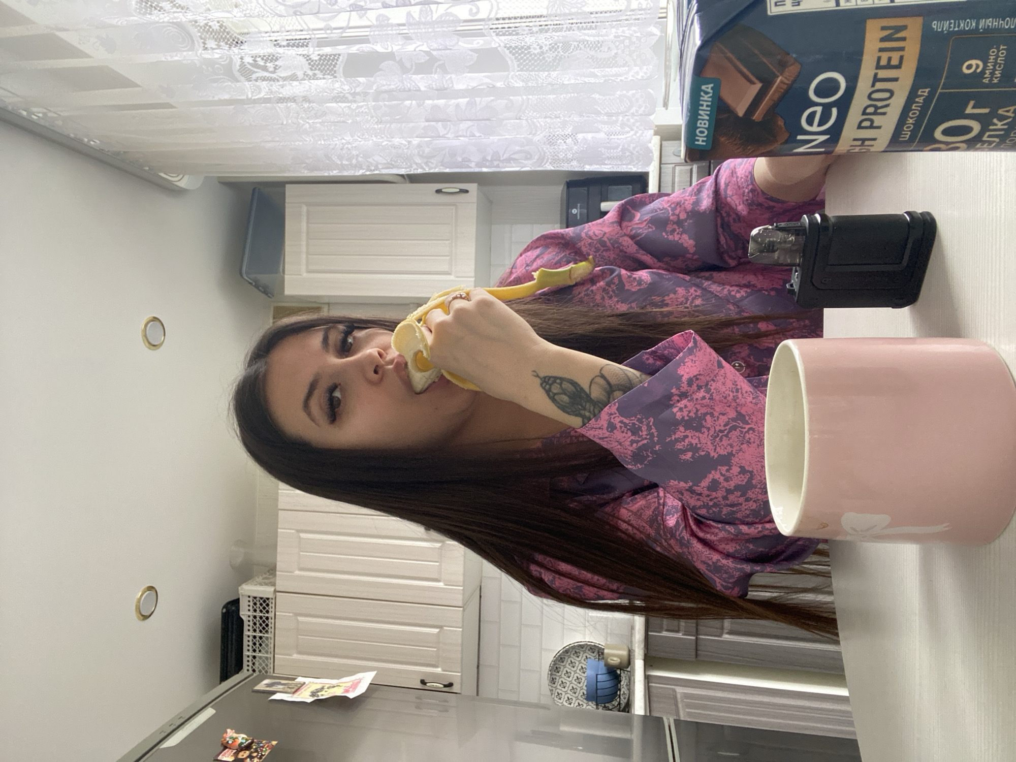 A woman with long brown hair is seated at a table, eating a banana. She is wearing a pink and purple floral pajama top. On the table are a pink mug, a black vape device, and a carton of "Neo High Protein" chocolate drink. The background shows a kitchen interior with white cabinets, white tiled walls, and a white lace curtain. The kitchen is located in Chaikovsky, Russia.