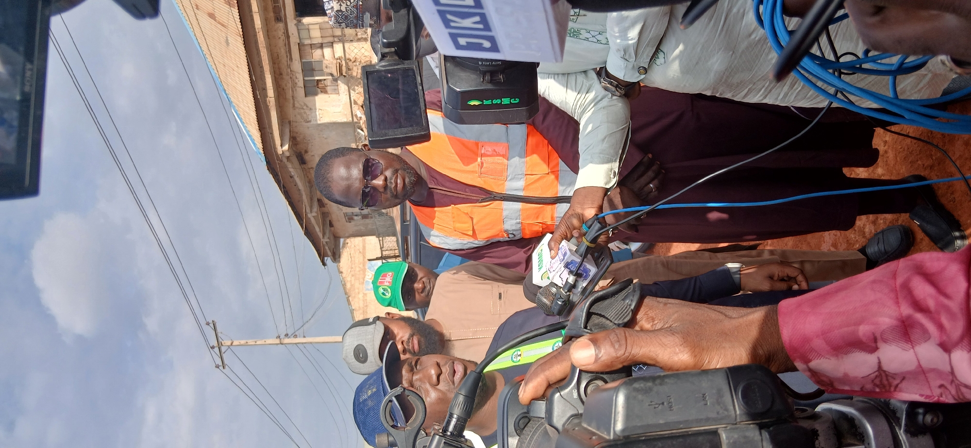 In Zaria, Nigeria, a man wearing an orange reflective vest and sunglasses, with a burgundy robe underneath, is being interviewed by reporters. He is surrounded by other men, some of whom are wearing caps. One reporter holds a microphone and camera, while another holds a second microphone with a logo. The background shows a building with a corrugated iron roof and a cloudy sky with telephone poles and wires. The ground is reddish-brown, suggesting a dirt or unpaved surface. The scene appears to be outdoors, possibly on a street or in a public area.