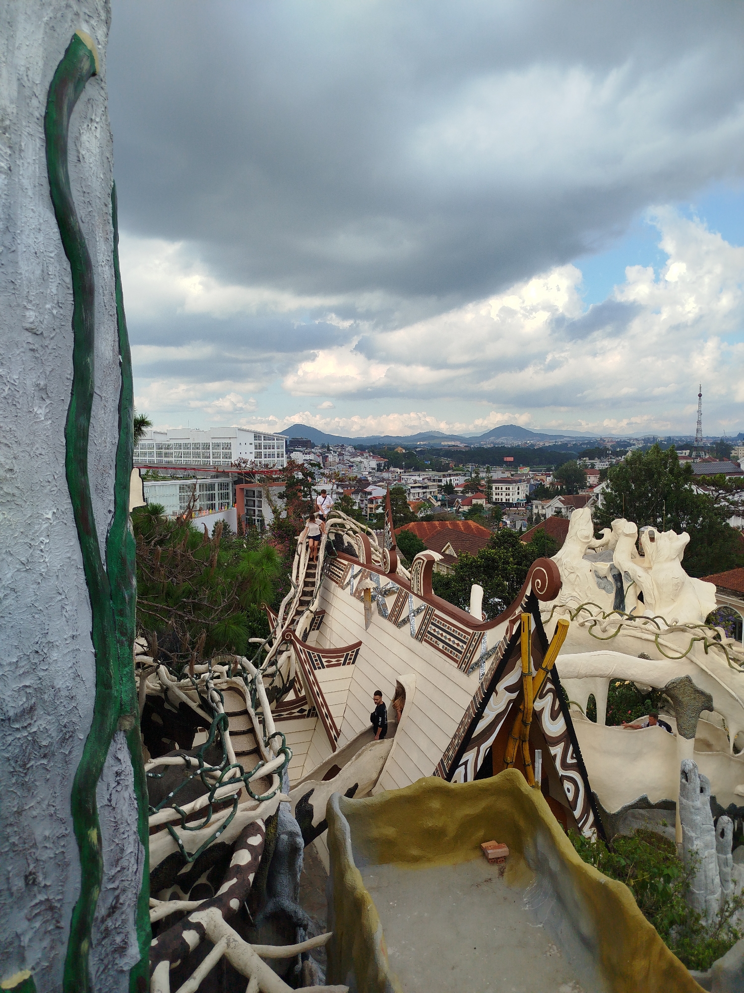 The image depicts a high-angle view within the Crazy House (Hang Nga guesthouse) in Da Lat, Vietnam. The architecture is whimsical and organic, resembling a tree or a network of interconnected structures with unconventional shapes.
The color palette is dominated by beige, brown, and gray, with accents of green and yellow. The buildings and structures are intricately decorated with geometric patterns and textures.
A child is visible on a bridge-like structure. In the background, the city of Da Lat unfolds, with buildings, trees, and distant hills visible under a sky with scattered, dark clouds. The overall impression is one of fantastical architecture within a more conventional urban landscape.
