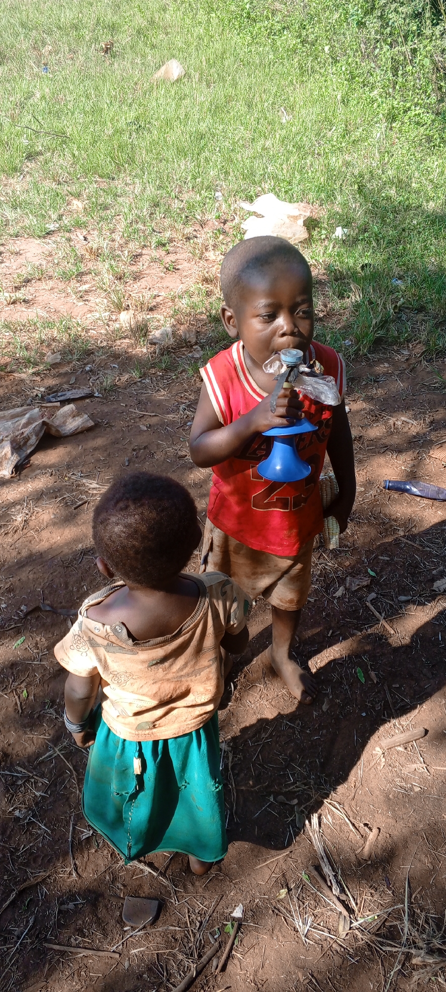 The photograph depicts two children in Ngalula, Democratic Republic of the Congo. One child, wearing a red sleeveless shirt and light-colored shorts, is standing and holding a makeshift drinking device made from a blue plastic funnel and a plastic bag. The child appears to be drinking from it. The second child, wearing a tan shirt and a green skirt, stands facing away from the camera. The ground is dirt with some scattered debris and sticks, and there is a patch of grassy vegetation in the background. The lighting suggests it is daytime.