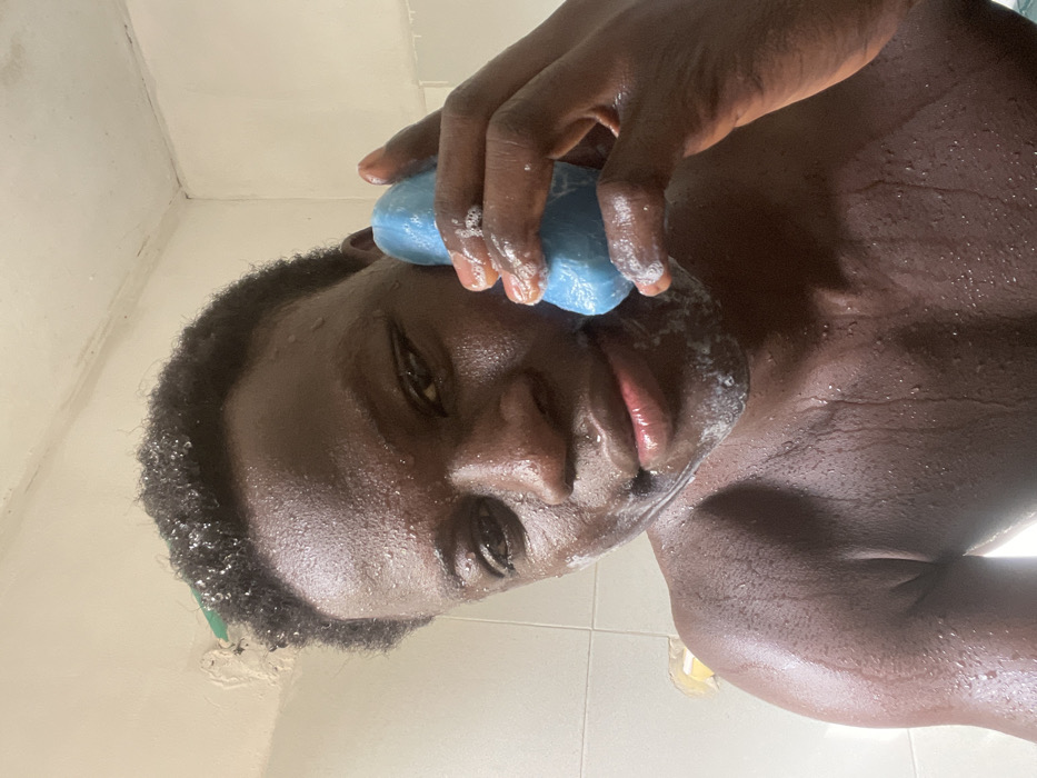 A dark-skinned individual, appearing male, is depicted in a close-up, chest-up shot within what appears to be a bathroom in Mosafejaw, Nigeria. The person has short, dark, curly hair, visibly wet and adorned with water droplets, similar to the skin across the face, neck, and upper torso. White soap suds are present on the chin and around the mouth area. The individual holds an irregularly shaped, solid blue bar of soap in their right hand, positioned against the right side of their face, near the cheek/jawline. Soap suds are also visible on the blue bar and the fingers of the right hand. The background consists of white ceramic tiles, with visible grout lines and some discoloration or dirt on the upper walls and ceiling edge, including what may be peeling paint or grime, and a small segment of a green object or pipe on the upper left. A small yellow object is partially visible on the lower left background tile. The person is looking directly forward, towards the camera.