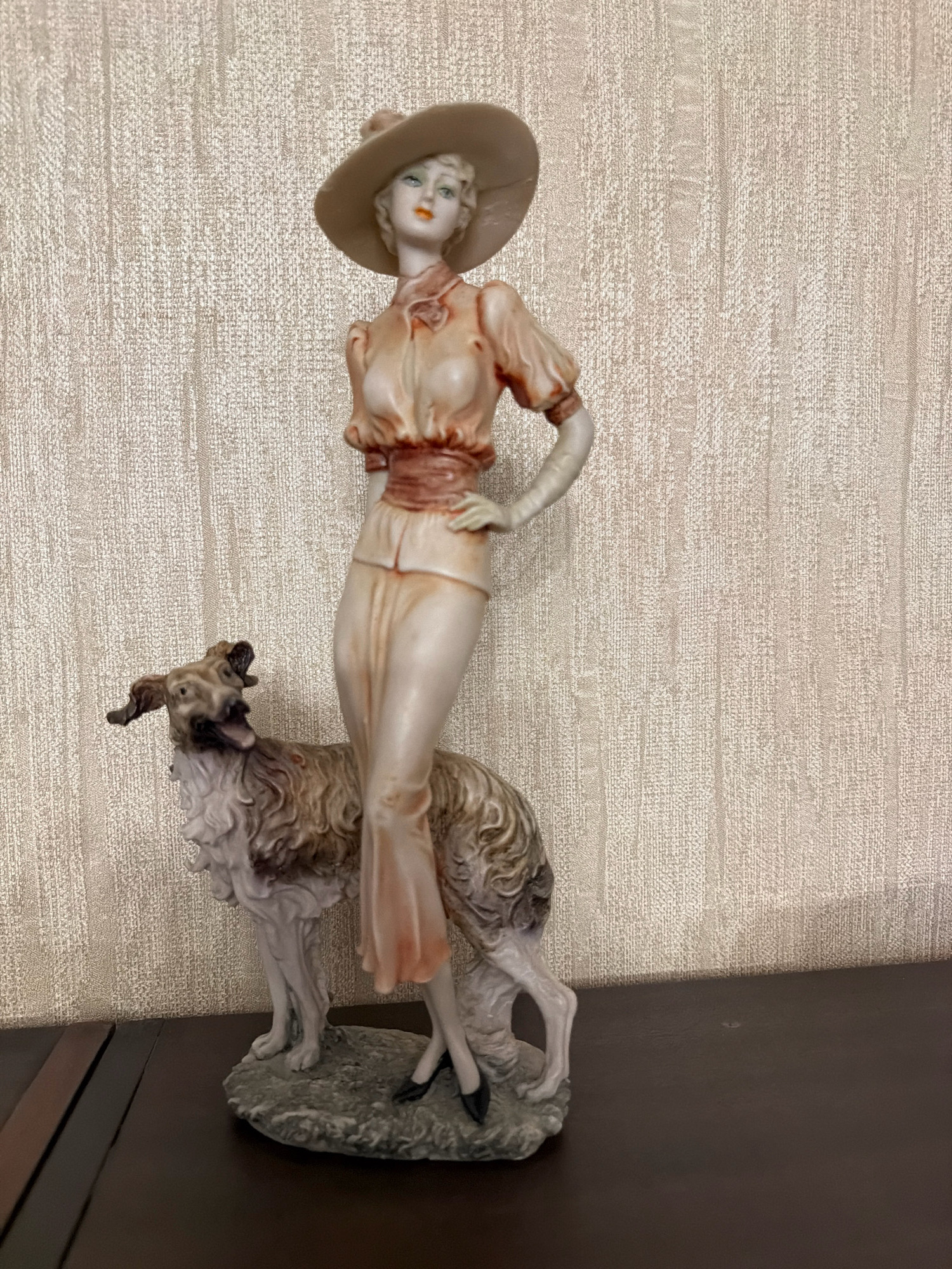 A figurine of a woman and a dog is displayed on a dark wooden surface, against a textured, light-colored wall. The woman, wearing a wide-brimmed hat and a peach-colored dress, stands with one hand on her hip. Her dress features puffed sleeves and a cinched waist with a reddish-brown sash. The dog, a long-haired breed, is positioned at the woman's side, with its mouth open as if barking. The figurine rests on a grey, rocky base. The scene is indoors, likely in a residential setting. The location is Sabunchu Raion, Azerbaijan.