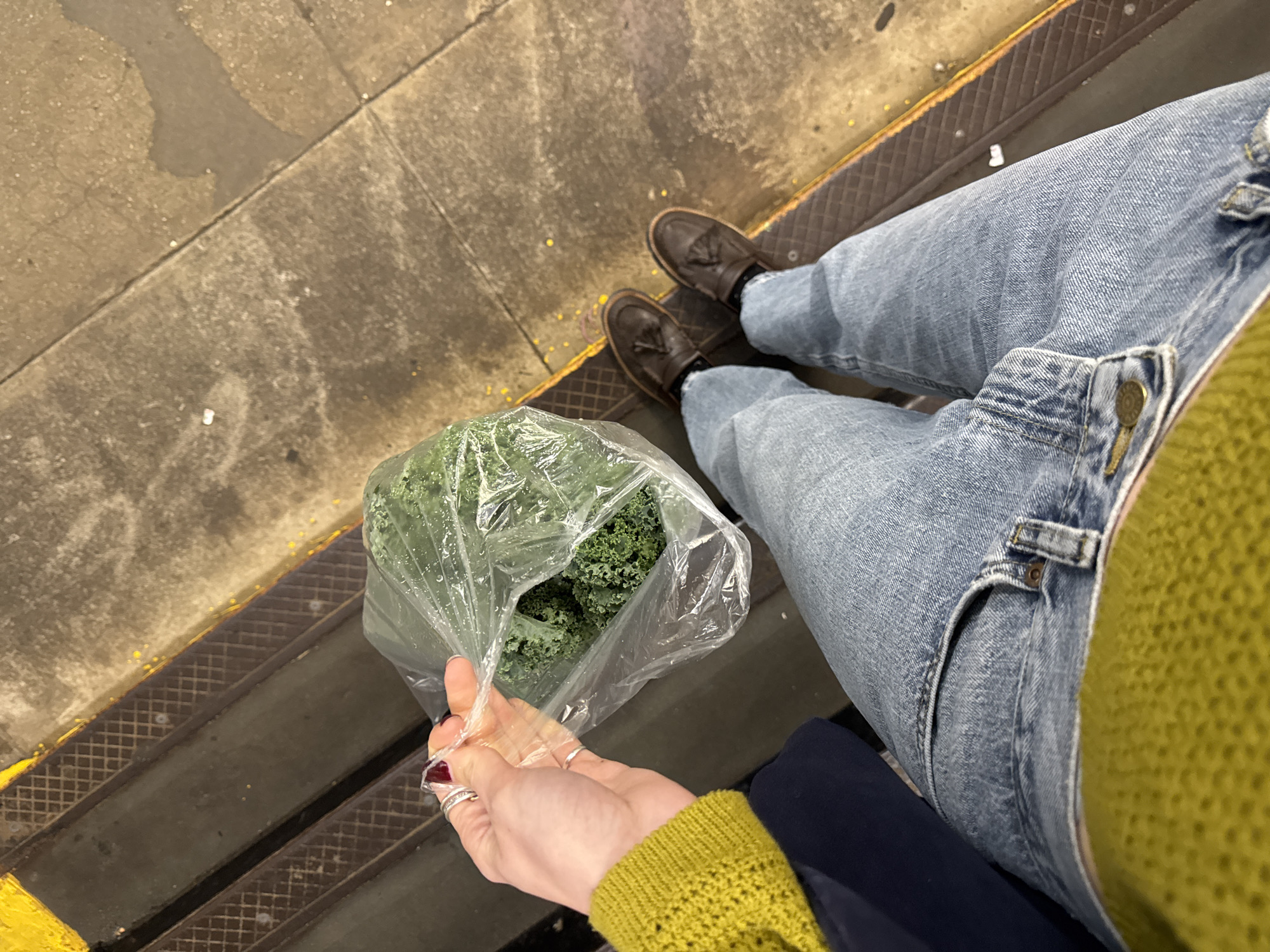 The photograph shows a person's-eye-view of someone standing on a New York City subway platform. The person is wearing blue jeans, brown tassel loafers, and a yellow knit sweater. In their hand, they are holding a plastic bag filled with kale. The subway platform has yellow safety treads and the ground is concrete. The picture captures the urban setting of a typical NYC subway station.