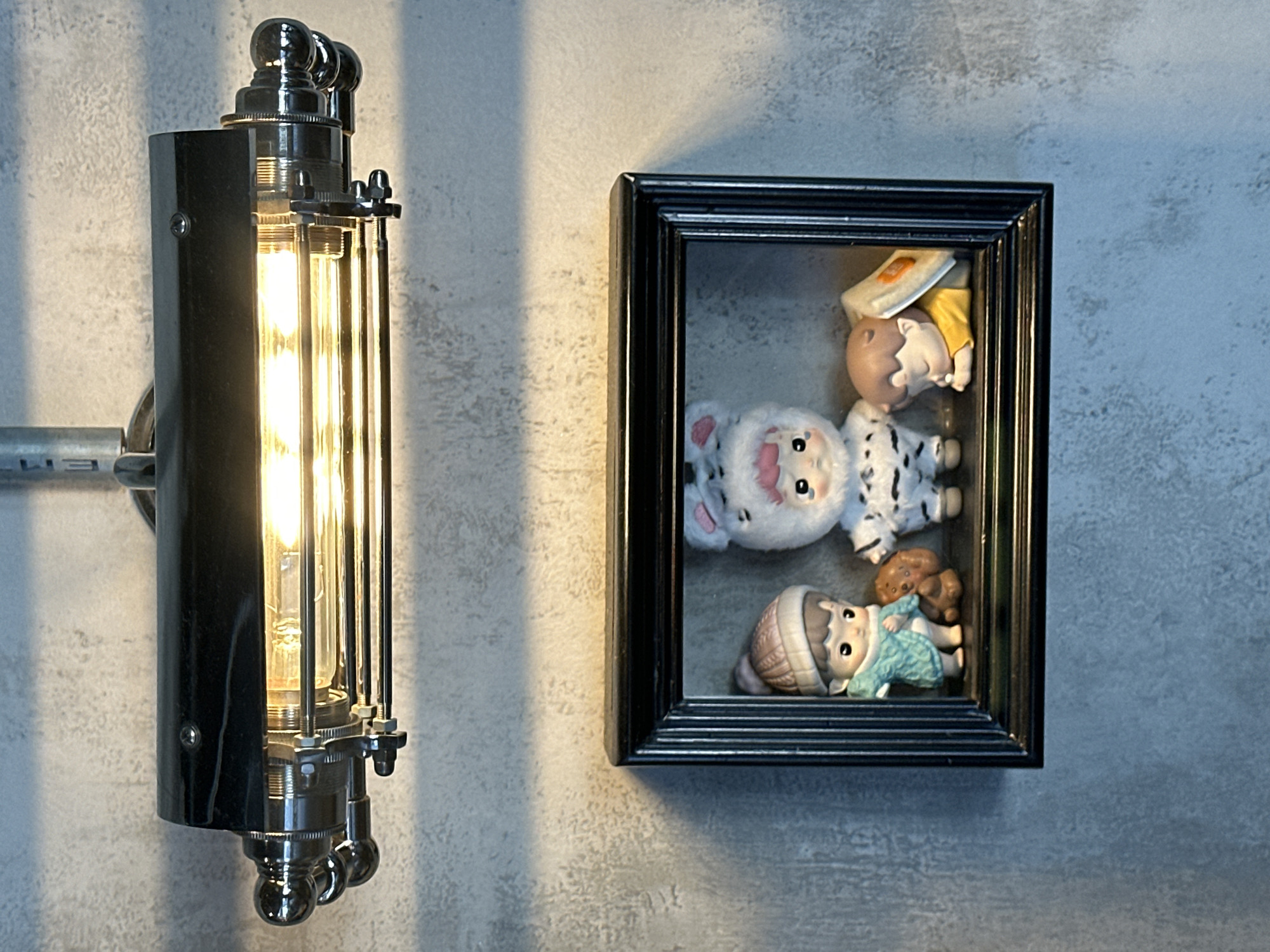 The image shows a close-up view of a framed display containing three figurines and a small stuffed toy, mounted on a textured grey wall. Above the frame, a vintage-style industrial light fixture with exposed Edison bulbs illuminates the scene.
The figurines, arranged from left to right, include:
1. A child wearing a teal knitted sweater and a pink beanie, holding a small brown teddy bear.
2. A child dressed in a fluffy white costume with cat or bunny ears and pink accents, featuring a black and white patterned outfit.
3. A child lying down, asleep, wearing a yellow shirt, with their head resting on a book or tablet.
The frame is black with a ribbed texture on its inner edge. The wall appears to be concrete or plaster with a noticeable texture. The lighting creates shadows on the wall, particularly from the light fixture.
This image does not provide any information about its location in Đà Nẵng, Vietnam.