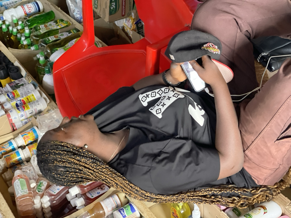 The image shows an adult female seated in an indoor commercial space, likely a shop or storage area, in Samaru, Nigeria. The subject has dark skin, long black and golden-brown micro braids, and is depicted in a profile view looking towards the right. She wears a black short-sleeved t-shirt featuring a large white 'V' logo with floral patterns, brown cargo pants, and a black shoe with a silver chain accent. A silver stud earring is visible on her right earlobe, and she wears a thin silver necklace and a dark bracelet on her left wrist. In her left hand, she holds a white smartphone connected by a white cable and a black baseball cap with a red brim and a yellow/red logo. She is seated on a red plastic chair, partially visible. The background features wooden or cardboard shelves densely stacked with various consumer goods, including multiple bottles of liquids (e.g., 'Hypo,' 'Viron,' green bottles of dish soap or similar products), a container of cotton buds, and a cardboard box labeled 'Sure Multipurpose Wipes.'
