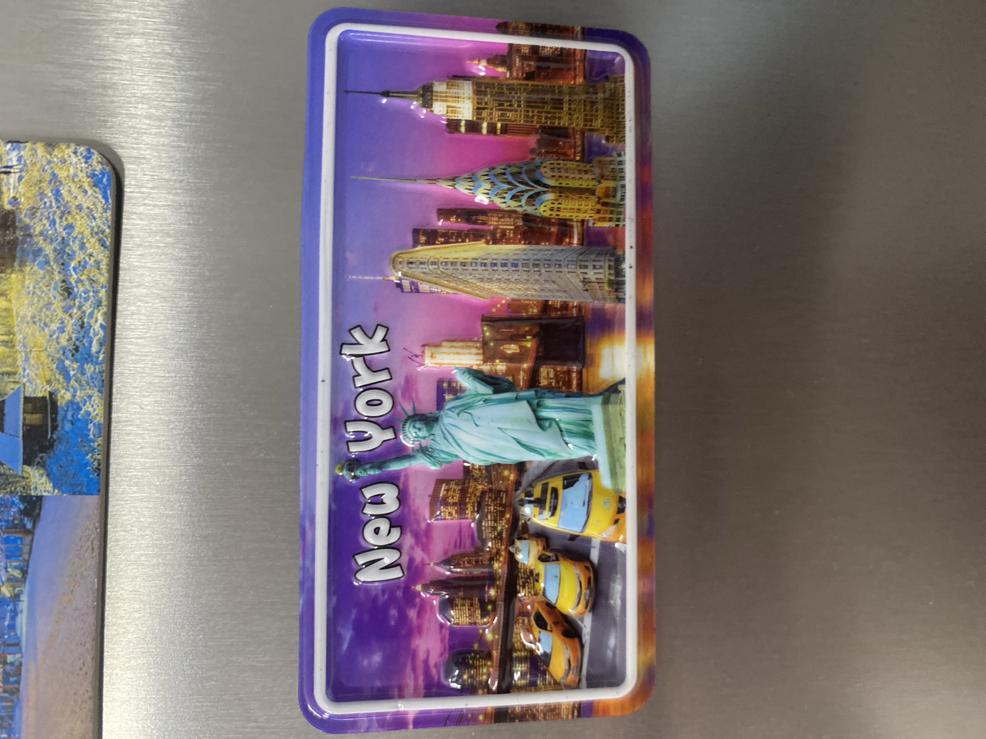 The image shows a rectangular magnet with a three-dimensional depiction of the New York City skyline. Prominently featured is the Statue of Liberty, rendered in a light blue-green hue, with its torch raised. Behind her, a vibrant cityscape unfolds, showcasing iconic skyscrapers like the Empire State Building and the Chrysler Building. The skyline is set against a dramatic purple and orange gradient sky, suggesting either a sunset or sunrise. Several yellow taxis are depicted on a street level below the Statue of Liberty. The text "New York" is visible on the magnet in large, raised white letters. The magnet is attached to a metallic surface, likely a refrigerator. To the left of the New York magnet, a portion of another magnet is visible, featuring a colorful, abstract design with blue and yellow elements, possibly depicting a landscape or artwork. The overall scene is static, with no discernible activity or event taking place beyond the representation of a New York scene. The location context provided is Łódź, Poland, however, the content of the magnet itself clearly depicts New York City.