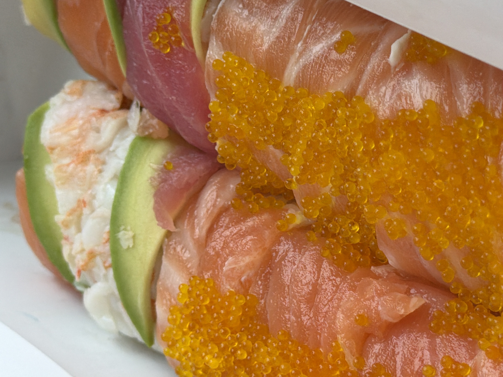 The image shows a close-up of two pieces of sushi. The sushi pieces are arranged in a stack within a white container. The bottom piece appears to be made with avocado slices, crab meat, and salmon. The top piece includes tuna, salmon, and orange roe. The scene appears to be indoors due to the controlled lighting and presentation. There are no discernible contextual clues to indicate the specific location in Tbilisi, Georgia.