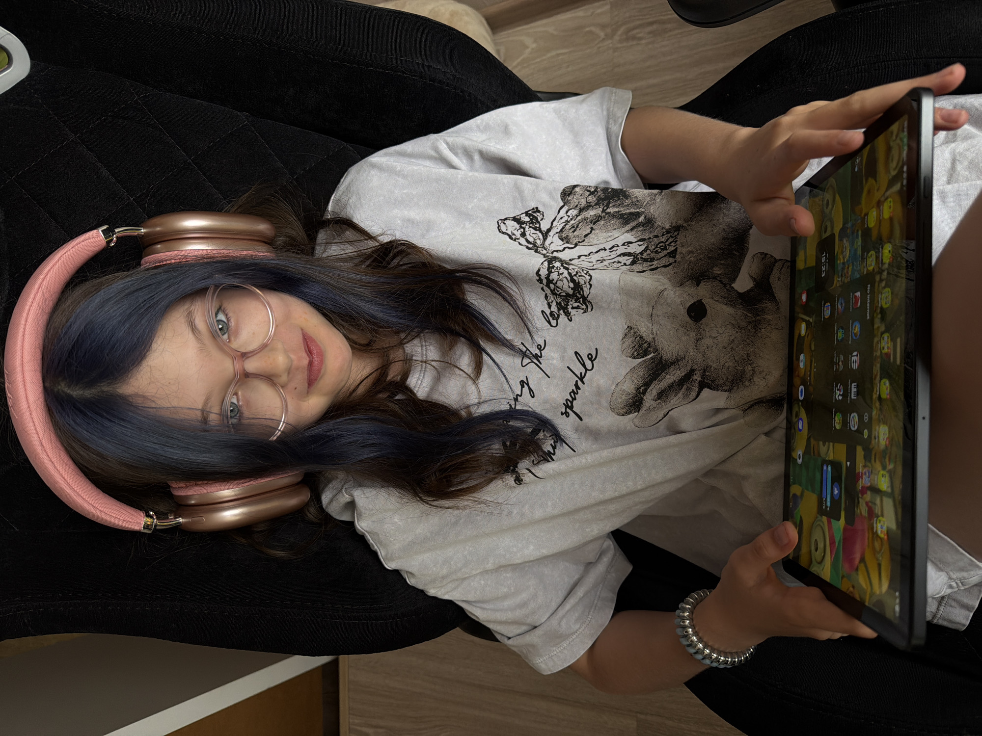 A young female, likely a child or teenager, is depicted lying on a dark, quilted surface, possibly a chair or couch, in an indoor setting. She is seen from a top-down perspective, looking directly at the camera with a slight smile or smirk. Her dark hair, with subtle blue streaks, frames her face, and she wears round, light-rimmed glasses over her light-colored eyes.
She is wearing large, pink/rose gold over-ear headphones and a light-colored, possibly tie-dyed or distressed, t-shirt with a graphic print. The print features what appears to be a butterfly and another animal, along with script text that reads "live the sparkle". On her right wrist, she wears a silver-colored coiled bracelet.
She holds a dark-framed tablet with both hands, positioned to her right. The tablet screen, angled towards the viewer, displays numerous app icons, a background image featuring yellow, Minions-like characters, and the digital time "15:23". A "No Internet" notification is also visible on the screen. The background of the scene includes glimpses of wooden or laminate flooring.
The overall impression is one of relaxed engagement, likely enjoying media or a game on the tablet. The time displayed suggests it is mid-afternoon. The location context provided is Birobidzhan, Russia.