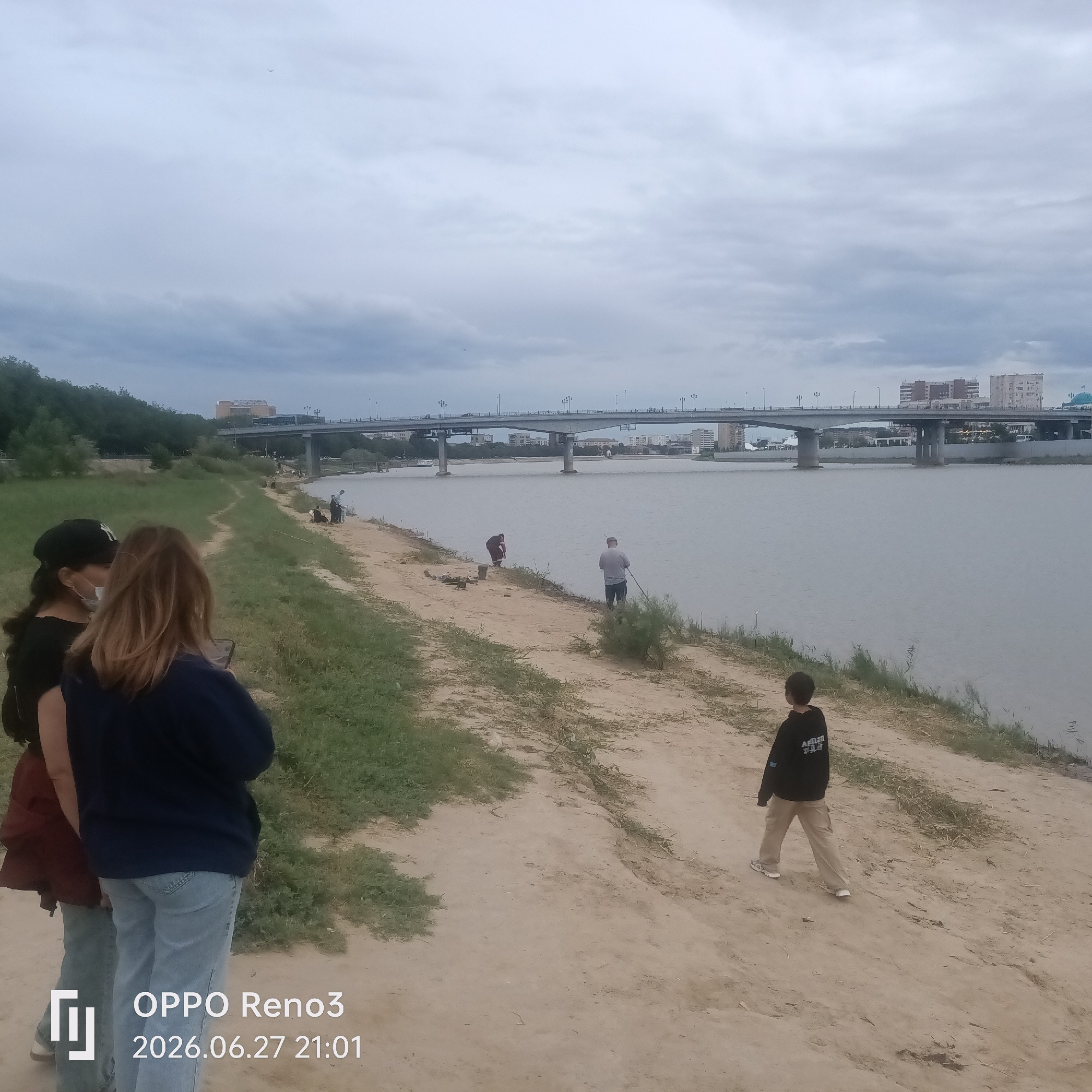 The image depicts a scenic riverside scene on an overcast day, likely in the late afternoon or early evening, given the diffused light. The setting appears to be Atyrau, Kazakhstan, with a wide river flowing calmly under a large bridge in the background. On the bridge, vehicles and streetlights are visible, suggesting an urban or semi-urban environment.
Along the sandy riverbank, several individuals are engaged in recreational activities. In the foreground, two women are standing, with one looking at her phone and the other appearing to take a photo or video. Further down the bank, a young boy in a black hoodie walks away from the camera, heading towards the river. Several other figures, seemingly fishing, are scattered along the bank near the water's edge, with fishing rods visible. The terrain is a mix of sandy soil and sparse green grass.
The sky is predominantly grey with cloudy formations, indicating fair weather without direct sunlight. The overall mood is relaxed and leisurely, with people enjoying the riverside environment. Visible text in the bottom left corner of the image indicates "OPPO Reno3" and the date "2026.06.27 21:01", which is a likely inaccurate future timestamp generated by the device.