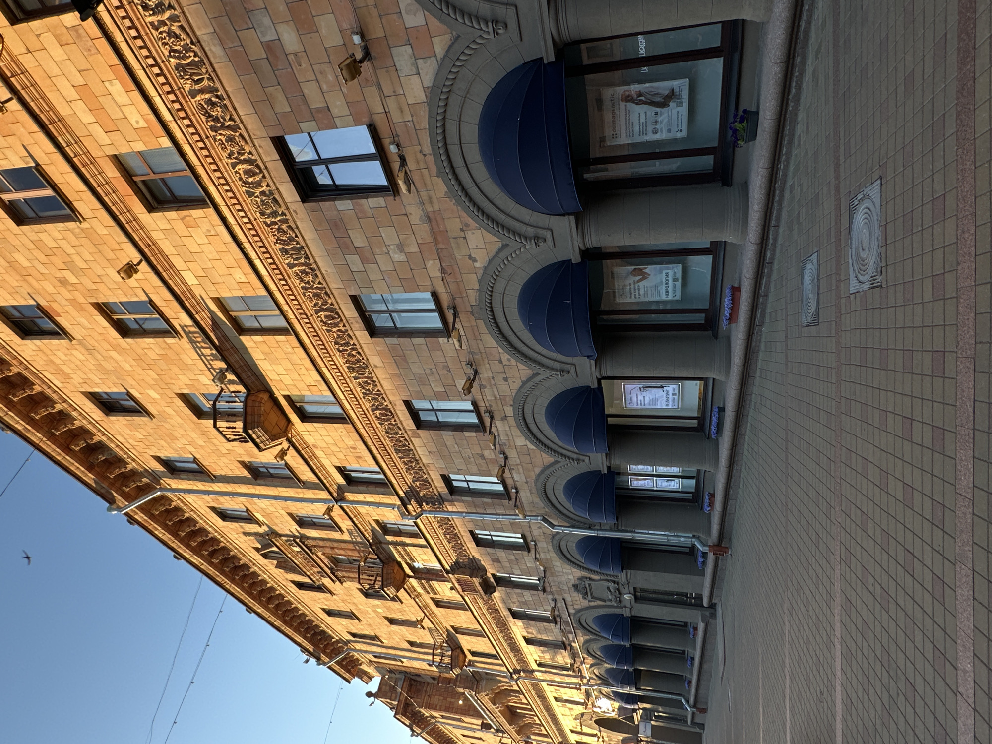 The image depicts a wide, sunlit street in Minsk, Belarus, dominated by an imposing, multi-story building constructed of warm-toned brick with decorative architectural elements. The building features numerous windows, some with dark frames and others with lighter ones, and several balconies jutting out from the upper floors. The lower facade of the building has a series of arched entrances, each covered by a dark blue awning, extending into the street. These entrances appear to be storefronts, with posters and signs visible through the glass.
The street itself is paved with patterned brickwork, and a walkway runs along the building's facade, lined with planters containing small purple flowers. The sky above is clear and blue, with a faint shadow of the building cast across the pavement, indicating the sun is low in the sky, likely in the late afternoon or early morning. A few power lines are strung across the sky. The overall impression is a well-maintained urban environment with a sense of historical architecture. There are no people visible in the image. The scene appears to be quiet and devoid of activity at the moment the photograph was taken.