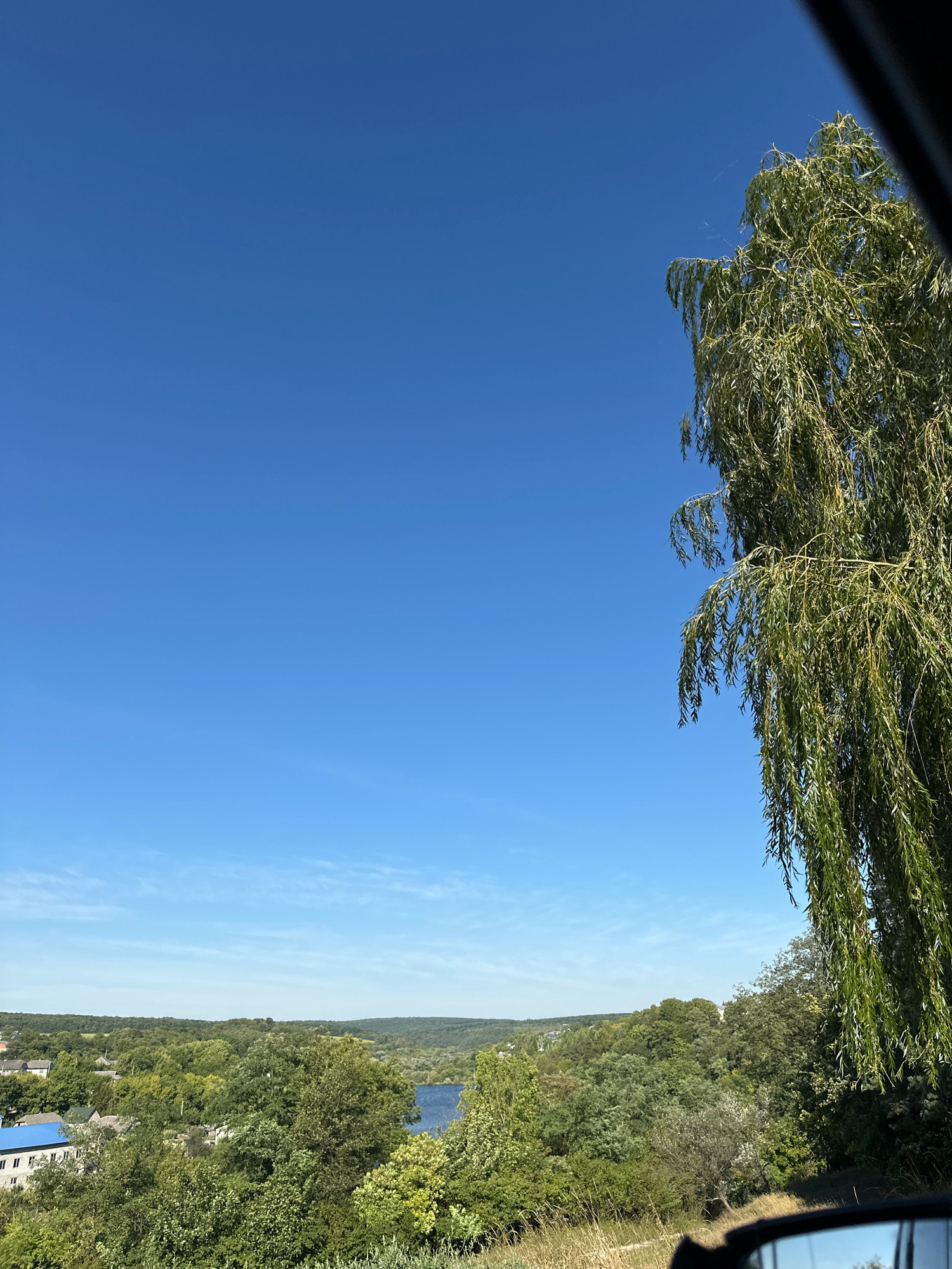 The image captures a scenic view of the city of Makiv, Ukraine, on a clear, sunny day. The foreground is dominated by the lush green foliage of a weeping willow tree, its branches gracefully cascading downwards. To the right, a portion of a car's side mirror is visible, indicating the photo was taken from inside a vehicle.
The midground reveals a picturesque landscape with rolling hills covered in dense, green forests. A serene body of water, likely a river or lake, is visible amidst the trees, reflecting the bright blue sky. In the distance, a small settlement with buildings featuring blue roofs can be seen nestled among the trees, adding a touch of human presence to the natural beauty. The sky is a brilliant, cloudless blue, suggesting fair weather. The overall scene is one of tranquility and natural charm, characteristic of a rural Ukrainian setting.