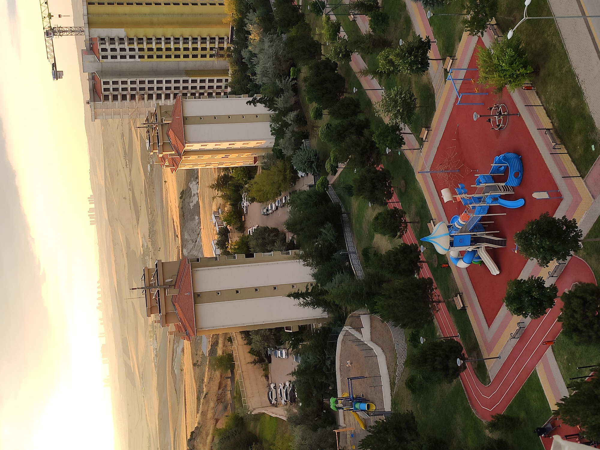 The image depicts a residential area in Etimesgut, Turkey, with a backdrop of a gently sloping landscape under a hazy, golden sky, likely at dawn or dusk. In the foreground, a children's playground is visible with bright blue and white play structures, including slides, swings, and a climbing net, all situated on a red rubberized safety surface. A running track with red and yellow lanes curves around the playground. Surrounding the playground and track are trees and park benches. Several apartment buildings are prominent in the middle ground, with one under construction, indicated by exposed framework and a large crane. A parking lot with several cars is visible near one of the completed buildings. In the distance, a city skyline is faintly visible on the horizon. The overall scene suggests a developing urban environment with amenities for residents.