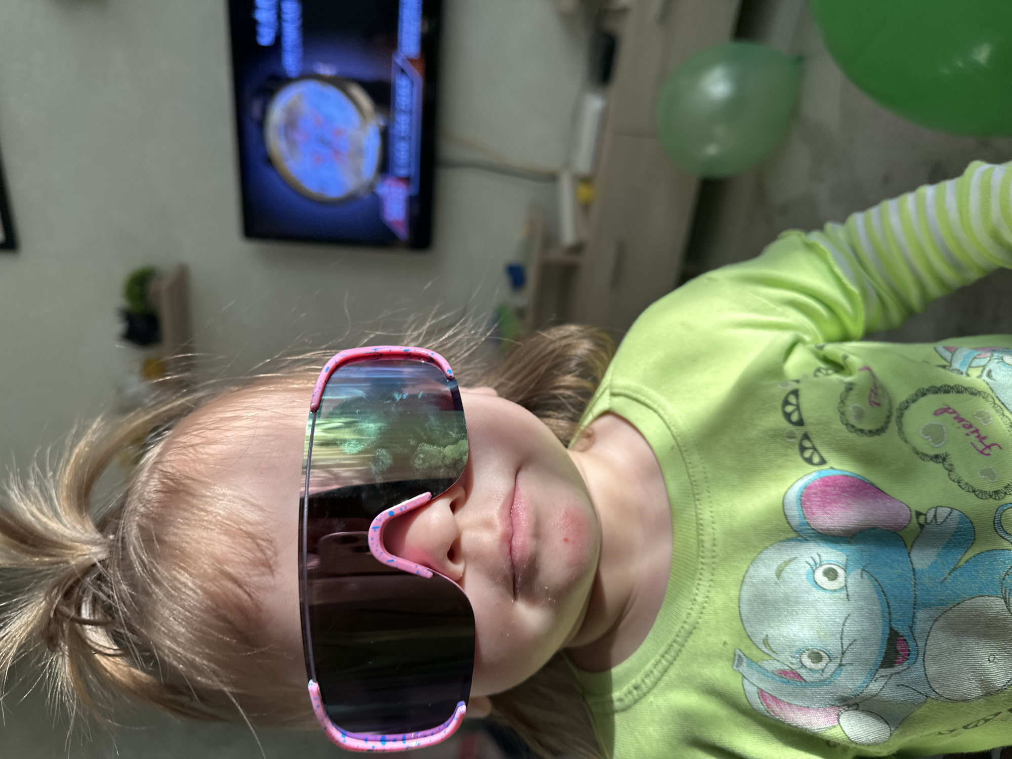 A young child is captured in a close-up, sideways portrait, set indoors in Uva, Russia. The child, likely a toddler or preschooler, has light blonde hair pulled back in a ponytail and is wearing oversized, reflective sunglasses with a pink frame accented with blue and pink specks. The lenses of the sunglasses show a distorted reflection of the surroundings, possibly including a window.
The child is wearing a light green long-sleeved t-shirt featuring a cartoon elephant design in blue, pink, and grey, along with other decorative elements. The word "Friend" is visibly embroidered or printed within a heart shape on the shirt's upper right side. A slight redness is noticeable on the child's upper lip area, extending towards the chin.
In the background, a television screen is visible, displaying what appears to be a graphic or map with prominent blue and red colors. Some indistinct white text or numbers are also on the screen. To the right of the child, partially visible, are two green balloons, suggesting a celebratory atmosphere. The lighting in the room appears bright, possibly from natural light, casting reflections.