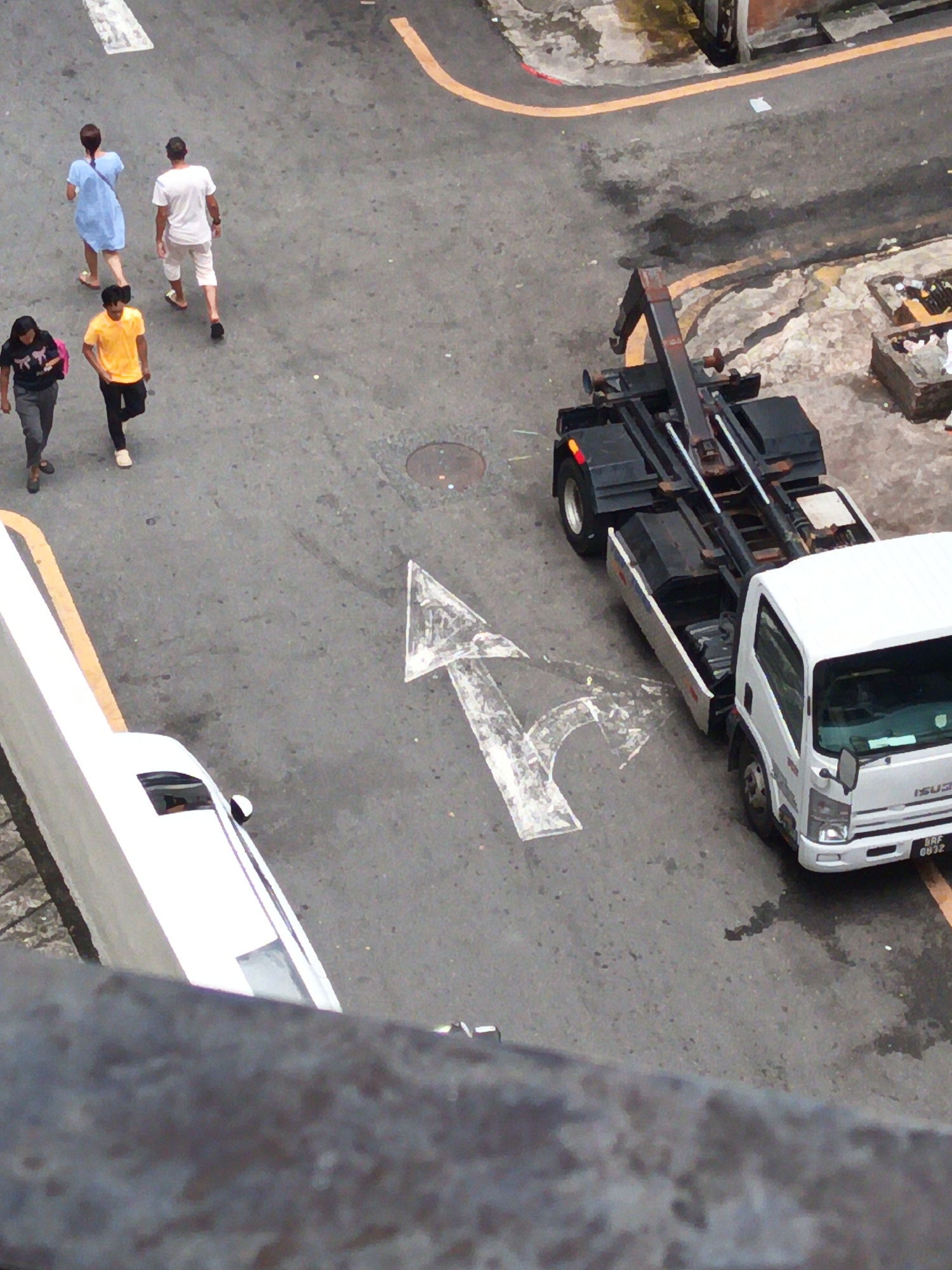An elevated, full-shot, eye-level view captures a street scene in Kuala Lumpur, Malaysia, on a cloudy day. Several individuals are walking on the asphalt. In the foreground, a white truck with a black crane attachment is parked on the right side of the road, facing away from the camera. The truck has a license plate that reads "BAF 6632". In the center of the image, a white arrow is painted on the road, indicating a turn. To the left, a white vehicle, possibly a car, is parked alongside the road. Further up the road, four pedestrians are visible. Two women, one wearing a blue dress and the other a black t-shirt, are walking away from the camera. Two men are also walking away from the camera; one is wearing a yellow t-shirt and the other a white t-shirt and white shorts. The road is marked with yellow lines and has various manhole covers and debris scattered on its surface. The overall impression is a typical urban street environment with vehicles and pedestrians engaged in their daily activities.