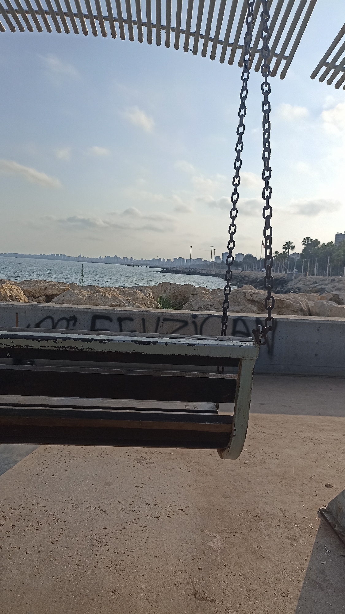 This media file captures a peaceful outdoor scene with a swing in the foreground, set against a backdrop of the sea and a distant city skyline. The swing, made of wood and suspended by metal chains, is the primary object. It hangs from a wooden structure with parallel slats, suggesting it's part of a park or recreational area.
The scene depicts a tranquil seascape with calm water stretching towards a horizon dotted with buildings. Large rocks form a barrier between the water and the paved area where the swing is located. Palm trees and more architectural structures are visible along the coastline, hinting at an urban or resort setting. The sky is a soft blue with scattered clouds, indicating fair weather. The lighting suggests it is either early morning or late afternoon.
Visible graffiti on a concrete barrier beneath the swing adds a touch of urban art to the natural and man-made elements. The graffiti appears to be in black lettering, but it's difficult to decipher fully. There's also a faint marking that looks like "ER" on the wooden slats of the swing itself.
The overall mood is one of serenity and quiet contemplation. The empty swing suggests a moment of pause, inviting relaxation and enjoyment of the view. The location, Yenişehir, Turkey, provides context for the architectural style and the presence of palm trees, consistent with a coastal Mediterranean environment. There are no people visible in the frame, emphasizing the stillness of the scene.
