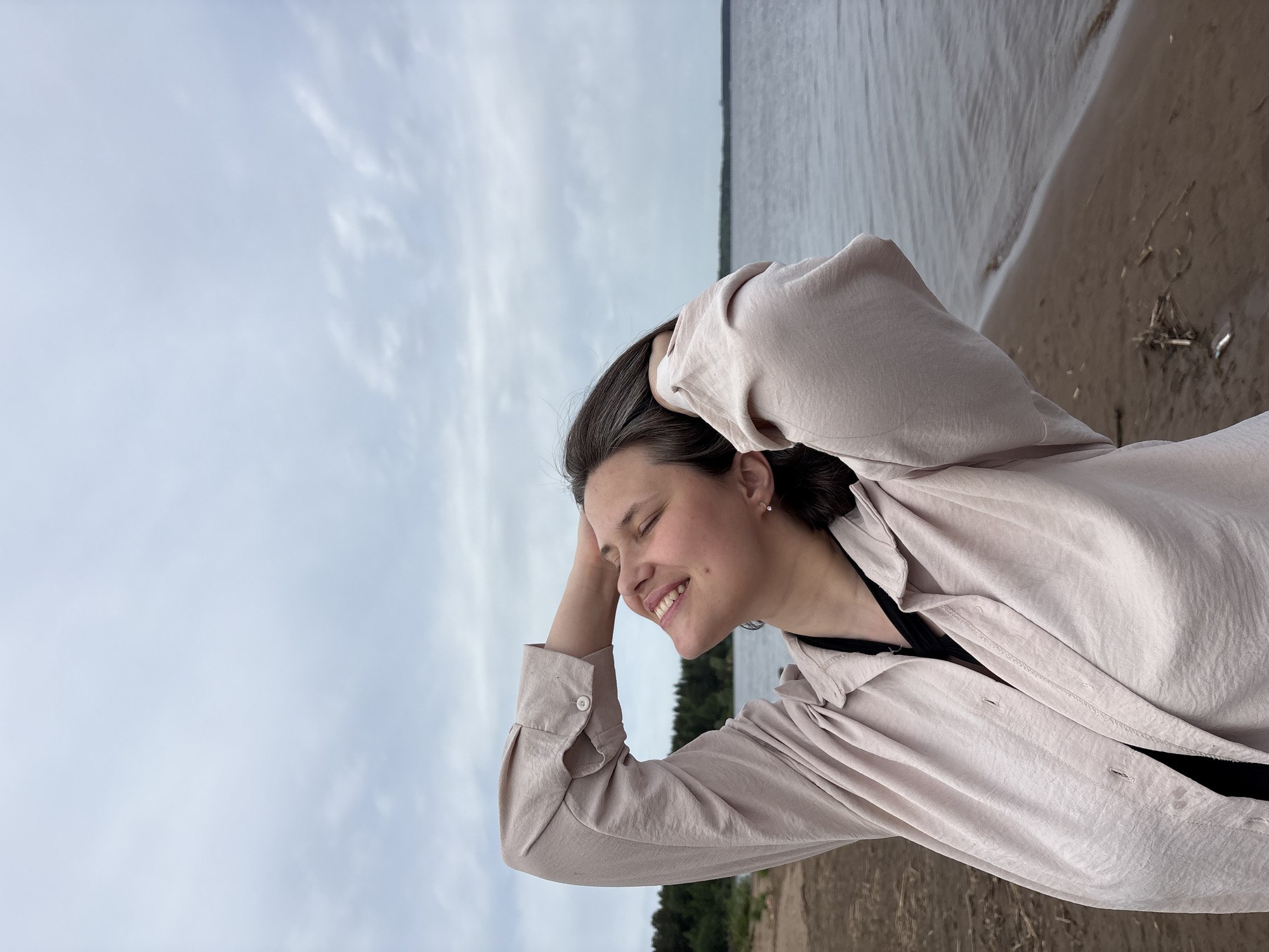 A young woman stands on a sandy beach in Chaikovsky, Russia. The sky is overcast. The woman is smiling with her eyes closed. She has her hands behind her head, wearing a light-colored button-down shirt over a black top. The edge of a body of water is in the background with trees on the far shore.