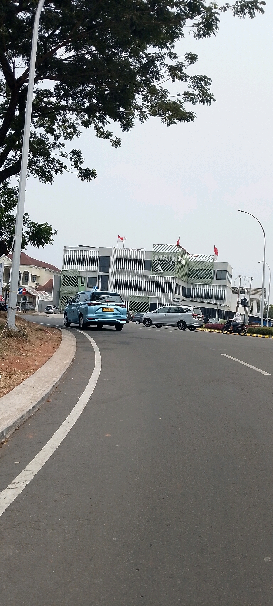 In Gunung Putri, Indonesia, a blue sedan, identified by its license plate B 1599 HII, drives on a paved road next to a concrete curb. Behind the sedan, a silver SUV drives in the same direction. Further down the road, a person rides a motorcycle. On the right side of the road, a modern building with white and green accents stands. The building's signage reads "MAIN STREET," and red and white flags are visible on its rooftop. The sky above is overcast, and a large tree with dark green leaves partially obscures the top left of the image. The scene depicts a typical street view in a commercial or residential area.