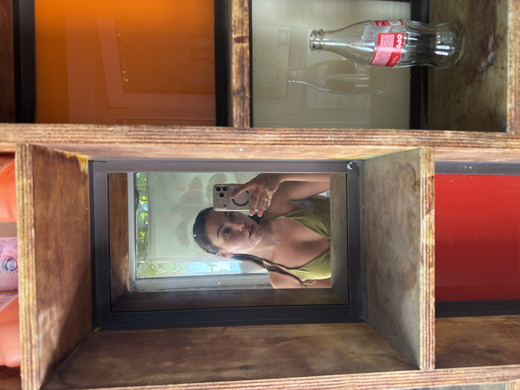 A young woman with dark hair pulled back is taking a mirror selfie in a rustic wooden shelving unit. She is wearing a green bikini top and a silver necklace. The shelving unit has multiple compartments, some containing decorative items like glass bottles and an orange-tinted panel. The background visible in the mirror suggests an outdoor setting with greenery. The image is taken indoors, in what appears to be a room with natural light.
