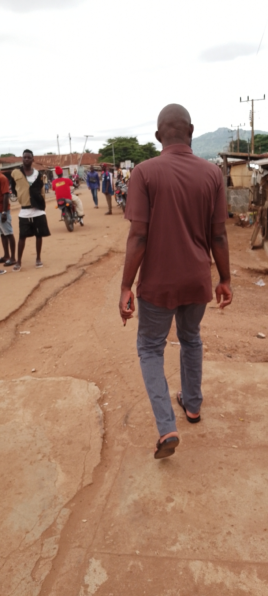 In Gboko, Nigeria, a bald Black man wearing a dark red polo shirt and grey pants walks away from the camera on a cracked, sandy road. He is holding a dark object in his right hand. In the background, several other people are visible, along with motorcycles, a building with a corrugated metal roof, and power lines against an overcast sky. To the left of the man, another Black man in a white tank top and black shorts stands looking towards the left. A person in a red shirt rides a motorcycle in the middle ground.