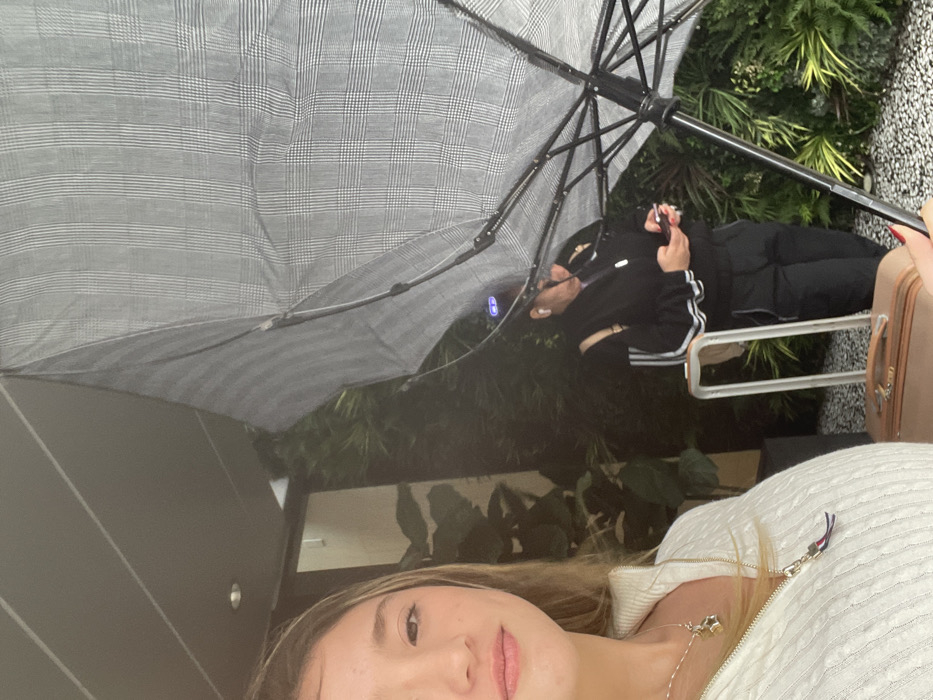 The image captures two women, one of whom is likely taking a selfie. She is blonde and wearing a white top and necklace. In the background, another woman wearing a black tracksuit is seated, looking at her phone under a plaid umbrella. They appear to be outdoors, with greenery visible in the background. A suitcase is partially visible on the right side of the frame.