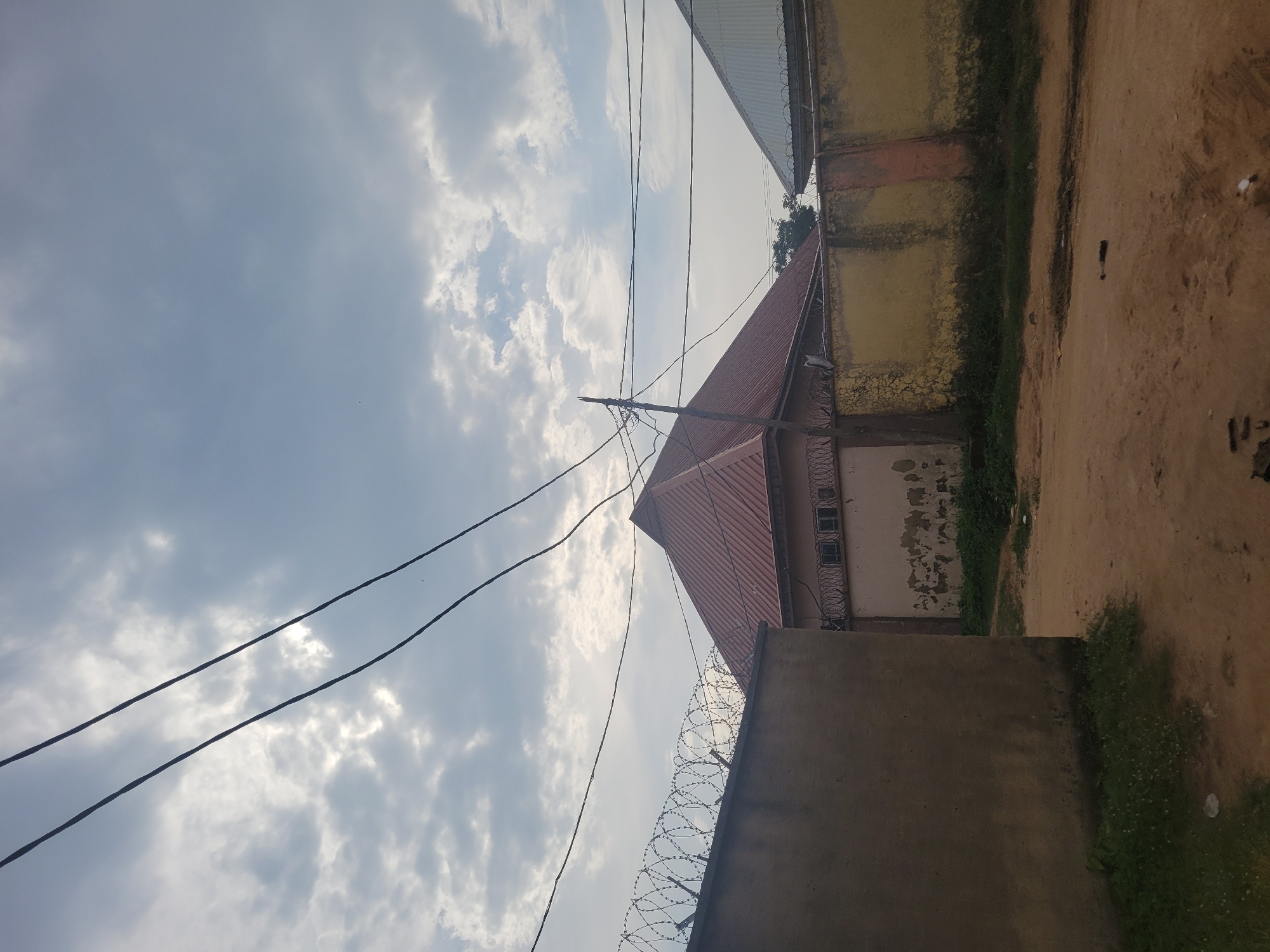 In the city of Suleja, Nigeria, an overcast sky with scattered clouds forms the backdrop for a view of residential buildings and surrounding walls. Two power lines traverse the upper portion of the frame. In the foreground, a dirt path leads past a rough, light-brown wall topped with razor wire on the left. To the right, a yellow-painted wall with patches of peeling paint and some orange staining runs along a property line. Behind the walls, a building with a red, pitched roof and a facade featuring several small, barred windows is visible. Another building with a similarly pitched, gray roof is partially obscured by the yellow wall. The environment appears to be a somewhat unkempt urban or semi-urban area, with sparse vegetation growing along the base of the walls and on the side of the path. The overall impression is one of a typical residential street in Suleja.