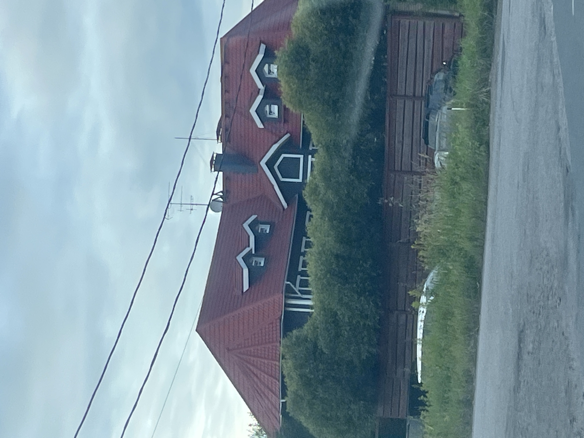 The image depicts a large, multi-story house with a red tiled roof located in Mar'ino, Russia. The house has dark colored walls, several dormer windows with white trim, and what appears to be a central gabled dormer with a diamond-shaped window. A dark chimney is visible on the roof. In front of the house, there is a dense line of green bushes, and behind that, a wooden fence. The foreground shows a grassy area with two vehicles partially obscured by the vegetation. One is a lighter-colored car, and the other is a darker SUV. The sky is overcast with clouds. Power lines are visible in the upper portion of the image.