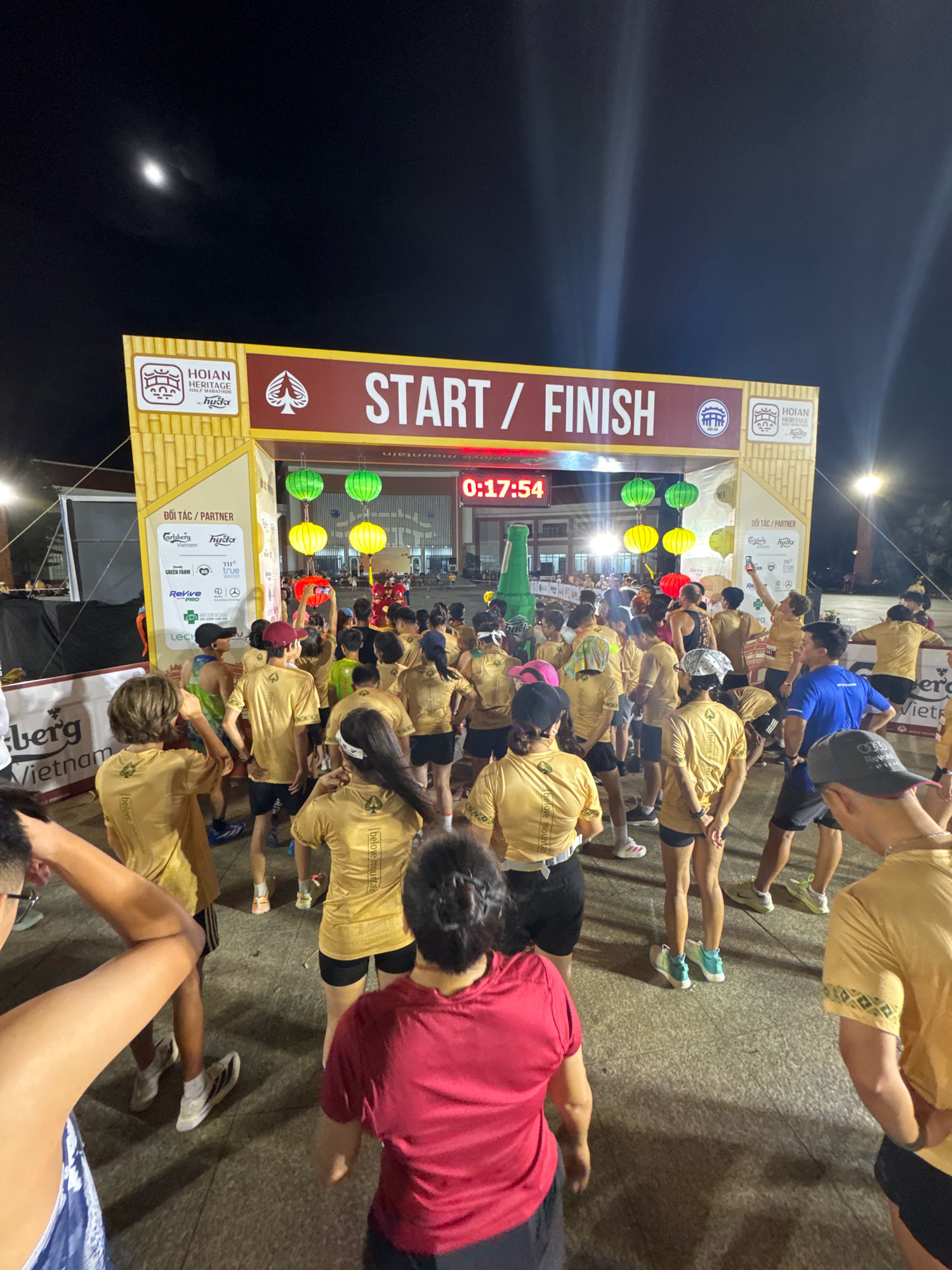 The image depicts the start/finish line of the Hoi An Heritage Half Marathon at night. A large archway with "START / FINISH" is prominently displayed. The archway is decorated with bamboo-themed panels and features logos and sponsors. Below the arch, a digital timer shows "0:17:54". Several runners, mostly wearing yellow t-shirts with a spade logo and the text "believe mountain", are gathered, awaiting the start of the race. The environment is illuminated by artificial lights, and colorful lanterns (green, yellow, and red) hang above the crowd. In the background, a large inflatable green bottle is visible. The scene suggests an outdoor event taking place on a paved surface. The moon is visible in the dark night sky.