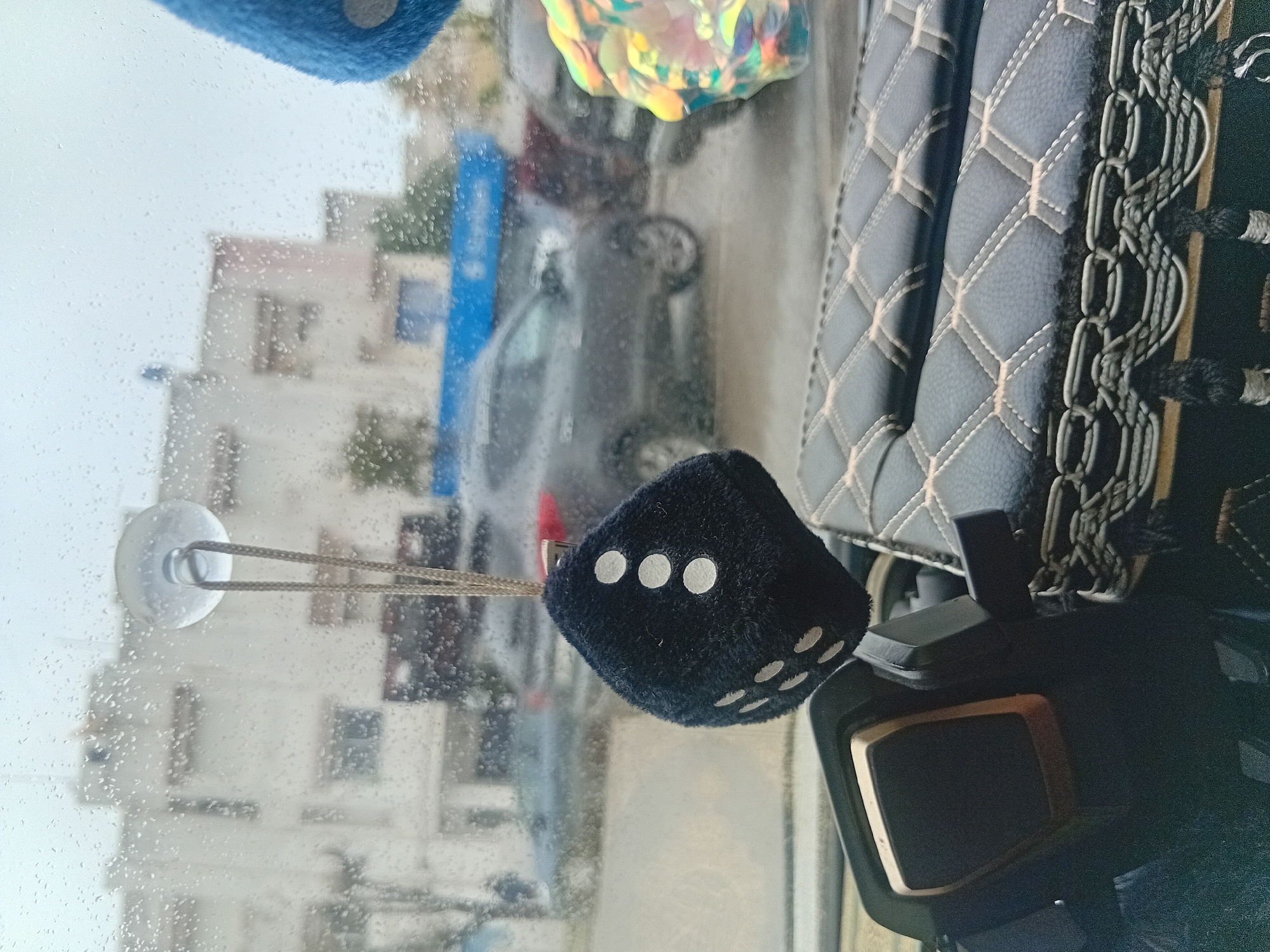 The image captures a view from inside a vehicle, looking out through a rain-splattered windshield into an urban or suburban setting in Martil, Morocco. The weather appears overcast and damp, with numerous water droplets visible across the glass, suggesting it is raining or has recently rained.
Inside the car, prominent objects include two fuzzy dice hanging from the windshield. A blue die is partially visible at the top, while a black die, showing faces with three and five white dots, hangs centrally from a white suction cup attached to the windshield via a cord. To the right, details of the car's interior are visible, including a grey or dark-colored quilted seat cover with light-colored stitching forming a diamond pattern, and a black and gold-trimmed car part, possibly part of the dashboard or a device. A colourful, iridescent, possibly holographic, ornament also hangs near the top of the windshield.
Through the wet glass, the blurred background reveals several light-colored buildings, likely residential, with visible windows. A dark-colored car is also discernable on the street or in a parking area, and a distinct vertical blue structure can be seen towards the left side of the exterior view. The scene is illuminated by daylight, but the overcast conditions create a soft, diffused light. There are no people visible, and no specific activity is taking place beyond observing the exterior scene from inside the car. No visible text is present in the image.