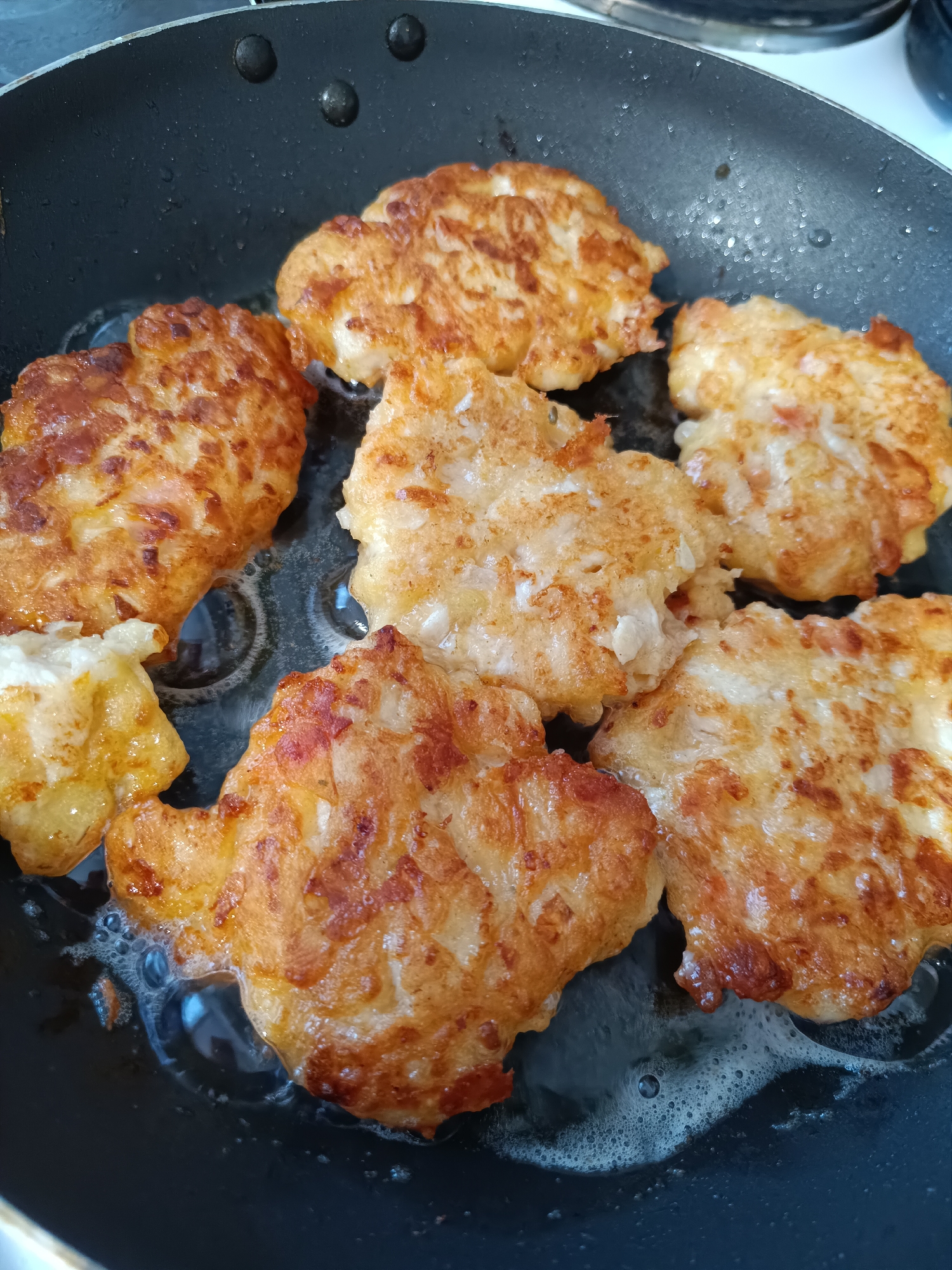 The image shows a frying pan with several golden-brown fritters cooking in oil. The pan is black, and the fritters have an irregular shape. Bubbles can be seen in the oil. The location is not discernible from the image.