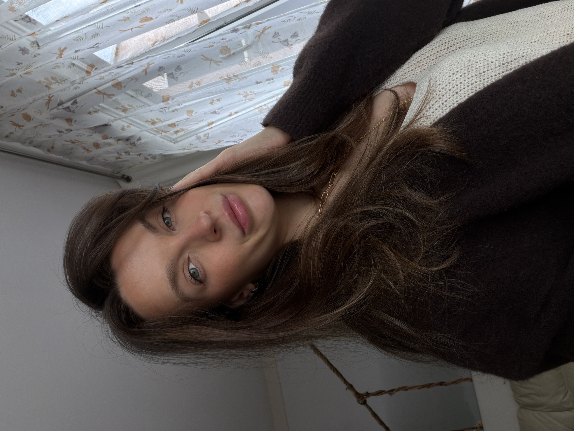 A young woman with long brown hair is pictured indoors. She is wearing a dark brown cardigan over a cream-colored knit top. She is looking directly at the camera, resting her hand near her temple. A gold chain is visible around her neck. Behind her, a window covered with a semi-transparent white curtain patterned with floral or botanical motifs can be seen. A white wall is visible on the lower left, while a lighter white surface is visible on the lower right. Without any other information the location in Budva is impossible to determine.