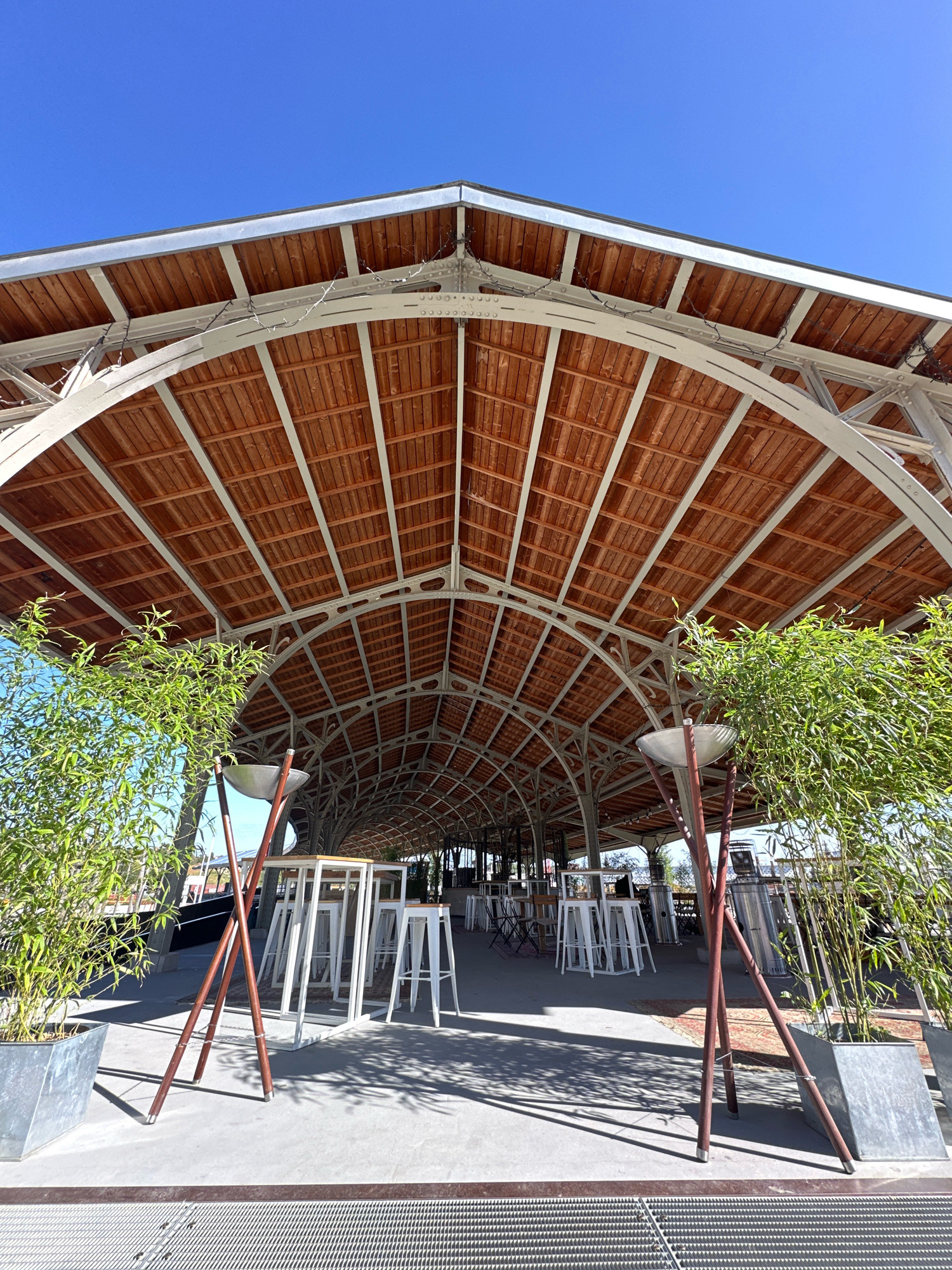 This image shows an outdoor seating area with a large, arched wooden roof structure supported by metal beams. The roof is lined with wooden planks, and string lights are visible along the metal framework. In the foreground, there are two tall planters containing bamboo plants. In front of the planters, there are tall, narrow tables with white stools, suggesting a bar or restaurant setting. The ground is paved with concrete, and shadows of the bamboo plants and tables are cast on it. The background shows more tables and chairs and appears to be under the extended roof. The sky is clear and blue.
The location is identified as Brussels, Belgium. Specifically, this is a view of the outdoor terrace at the Brussels Agriculural Hall (Halle agriculturale de Brussels).