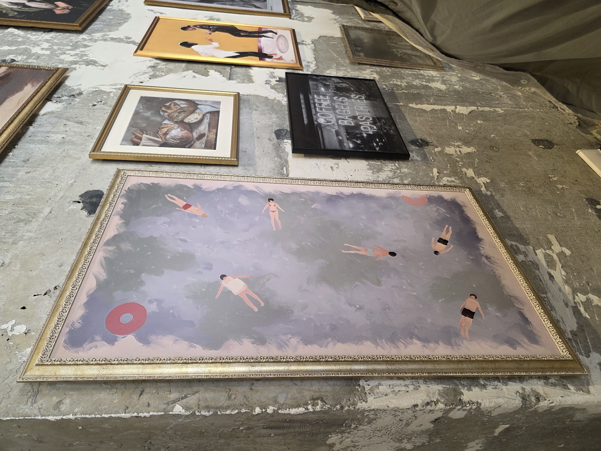 The image displays a collection of framed artwork adorning a distressed concrete wall. The primary artwork, a large vertical painting with a ornate gold frame, features a scene of figures floating in water. Several smaller framed pieces are also visible, including a still life of bread, a depiction of two figures in yellow and black, and a black and white image of neon signs reading "COFFEE BAGELS PASTRIES". The wall itself shows signs of age and wear, with peeling paint and textured surfaces. The lighting appears to be artificial, casting shadows and highlighting the artwork. The context of Khatay Raion, Azerbaijan is not evident from the visual information provided in the image.