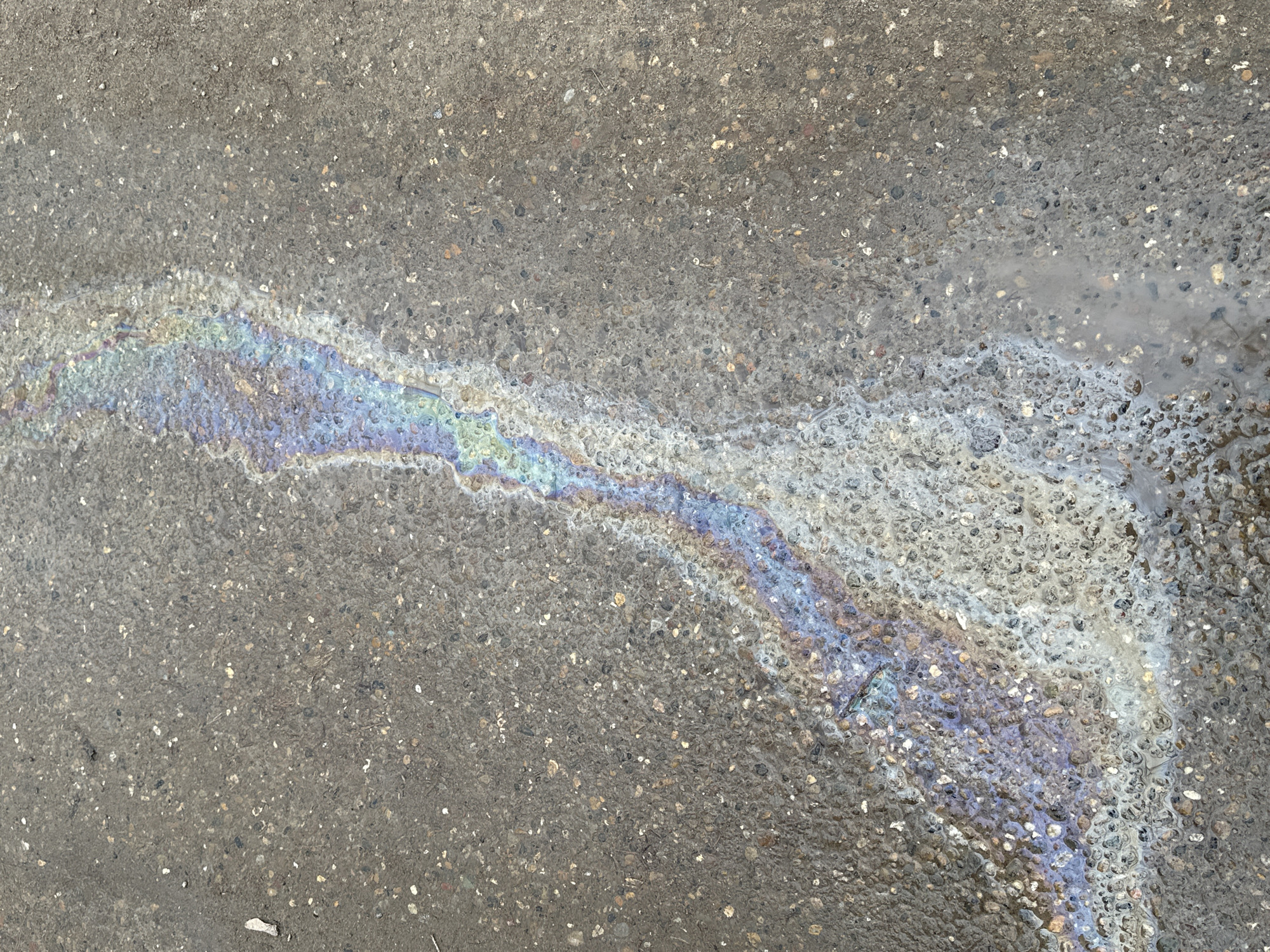 The scene depicts a close-up, high-angle view of a section of asphalt pavement in Chaikovsky, Russia. A distinct, elongated iridescent streak, characteristic of an oil or fuel spill on a thin layer of water, traverses the surface from the upper left quadrant diagonally towards the lower right. The spill exhibits a spectrum of colors including blues, greens, yellows, oranges, and purples. The asphalt itself is dark grey, coarse-grained, and appears damp, with embedded and scattered small aggregate particles. Minor light-colored debris and a slender, dark fragment of organic material (possibly a twig) are visible on the asphalt surface, primarily outside the most intense iridescent area. No individuals, actions, or interactions are present in the frame. The environment is an unidentifiable outdoor paved area.