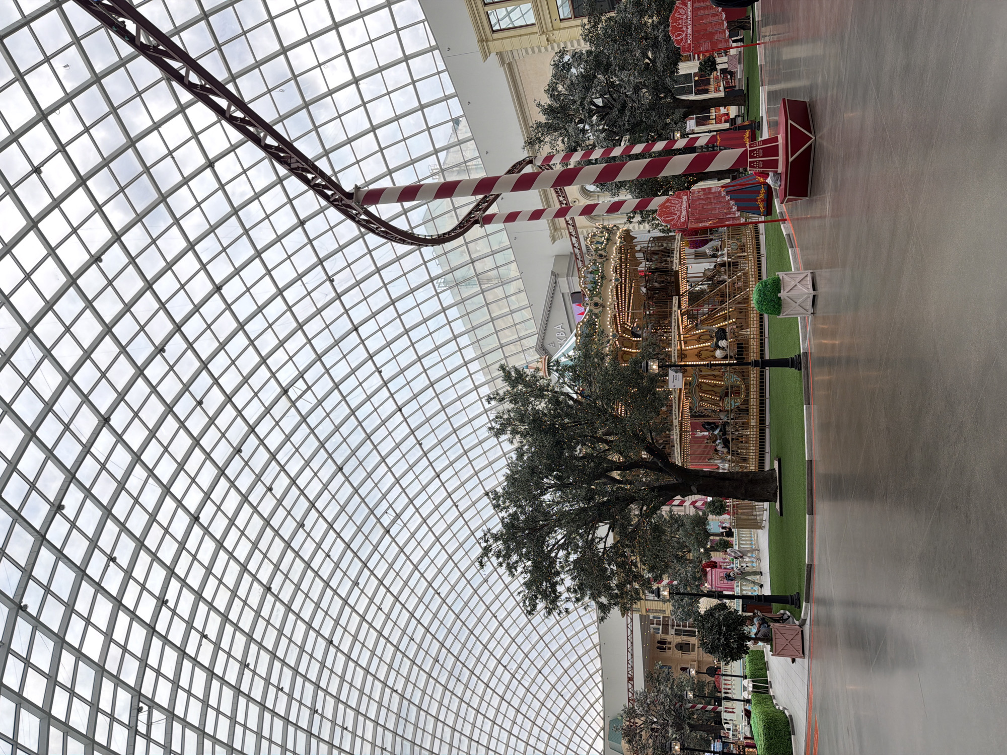 An indoor amusement park with a large glass-paneled domed ceiling is depicted. In the center of the frame, a traditional carousel with various animals is set up, adorned with lights and decorative elements. To the right of the carousel, a roller coaster track curves upwards, supported by large, red and white striped pillars. Several artificial trees with dense foliage are strategically placed throughout the space, adding to the park's ambiance. The ground is paved with a smooth, reflective material, and there are some green landscaped areas with hedges and grass. A few individuals are visible in the background, walking around or sitting on benches. The architecture of the surrounding buildings, visible beyond the amusement park elements, suggests a shopping mall or entertainment complex.