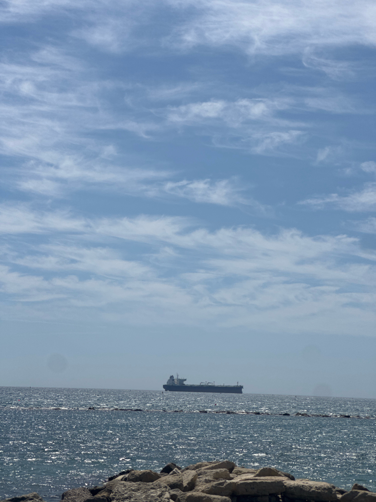 The image captures a serene seascape under a bright, partly cloudy sky. In the distance, a large cargo ship is visible on the horizon, its dark silhouette contrasting with the shimmering blue sea. The water is choppy, with sunlight glinting off the surface, creating a dazzling effect. In the foreground, a rocky shoreline with large, grey stones forms a natural barrier against the water. The sky is a pale blue, adorned with wispy white clouds that stretch across the frame, suggesting a gentle breeze. There are no people or animals visible in the frame, and no specific landmarks of Germasogeia Municipality, Cyprus are identifiable. The overall impression is one of a peaceful, expansive view of the sea.