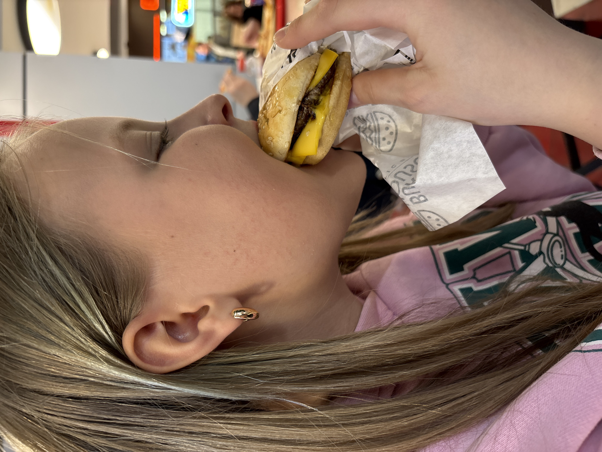 A young woman with blonde hair is holding and biting into a small cheeseburger wrapped in white paper. She is wearing a pink and white long-sleeved shirt and a gold earring. The background is blurred, but suggests an interior setting with light-colored walls and some other people in the distance. The restaurant location is in the city of Izhevsk, Russia.