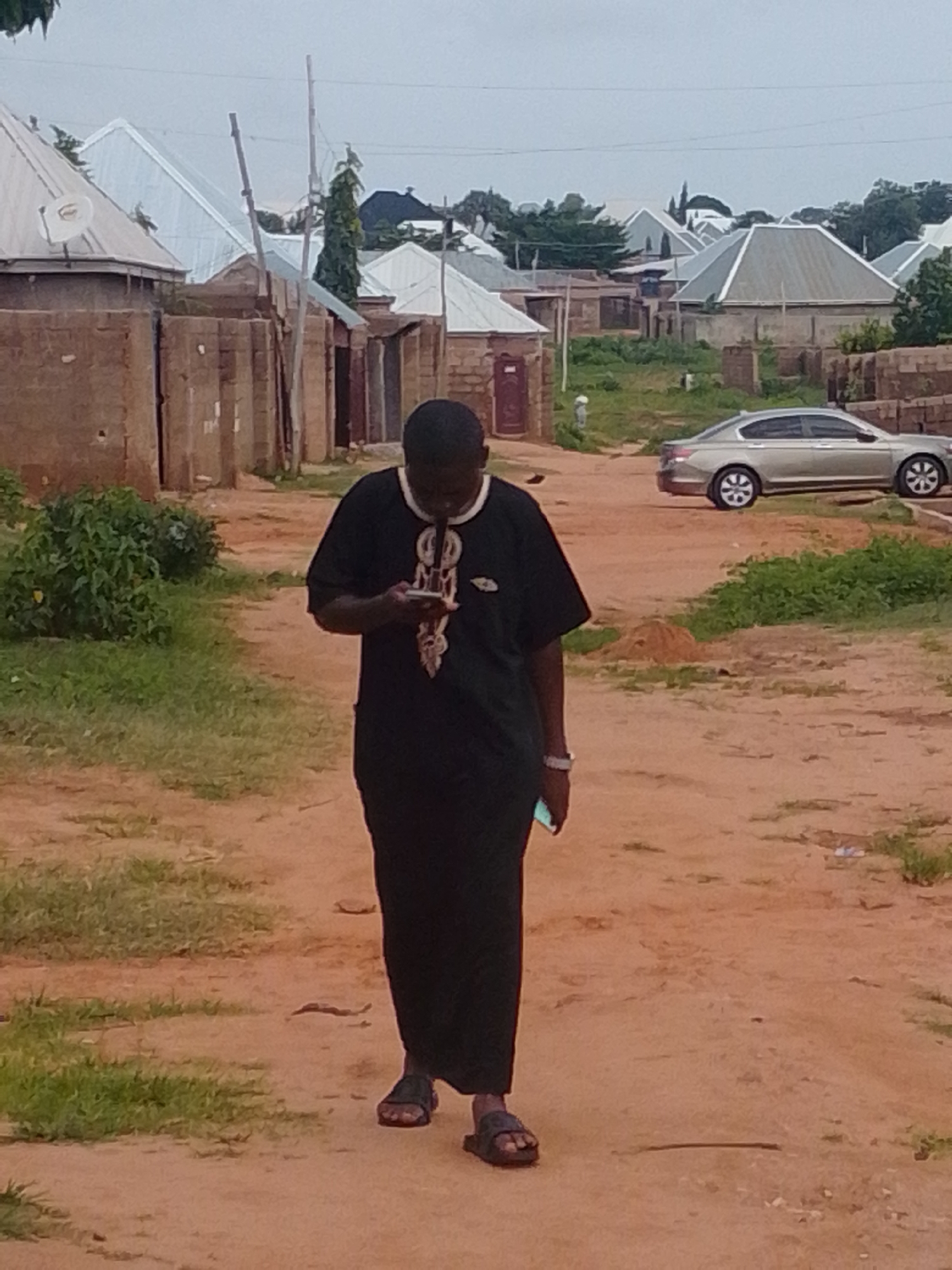 A medium shot captures a Black male wearing a long, black traditional garment with white embroidery on the chest, walking away from the viewer on a dirt road in the city of Gombe, Nigeria. The man is looking down at his phone. In the background, several buildings with corrugated metal roofs are visible, along with some trees and a silver sedan parked on the right side of the road. The sky is overcast. The ground is a reddish-brown dirt path with patches of dry grass on either side.