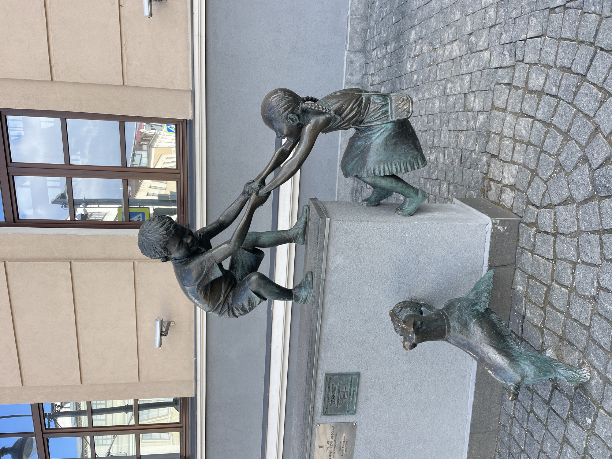 A bronze sculpture in Kazan, Russia, depicts a scene of childhood play and friendship. Two children are engaged in an activity, with a girl, wearing braids and a dress with a satchel, helping a boy to climb onto a low stone ledge or platform. She is bent over, extending her hands to pull him up, while the boy, in shorts, reaches out to her, seemingly in mid-climb. Below them, a small dog with perked ears looks up, observing the interaction.
The sculpture is set outdoors, against the light-colored facade of a building. Large windows on the building reflect the bright blue sky with some clouds, as well as reflections of other architectural structures. The ground is paved with grey cobblestones, arranged in a fan-like pattern in the foreground.
Two plaques are visible on the wall next to the sculpture's base. The upper plaque, in Russian, reads "ДОВЕРИЕ" (DOVERIE), meaning "TRUST," which is likely the name of the sculpture. The lower plaque mentions "ЛУЧШАЯ АРХИТЕКТУРНАЯ ФОРМА" (BEST ARCHITECTURAL FORM) from a "гра-конкурса 2016 года" (competition of 2016 year) and credits the "скульптор Альтаф Валиуллин" (sculptor Altaf Valiullin). The scene is illuminated by clear daylight.