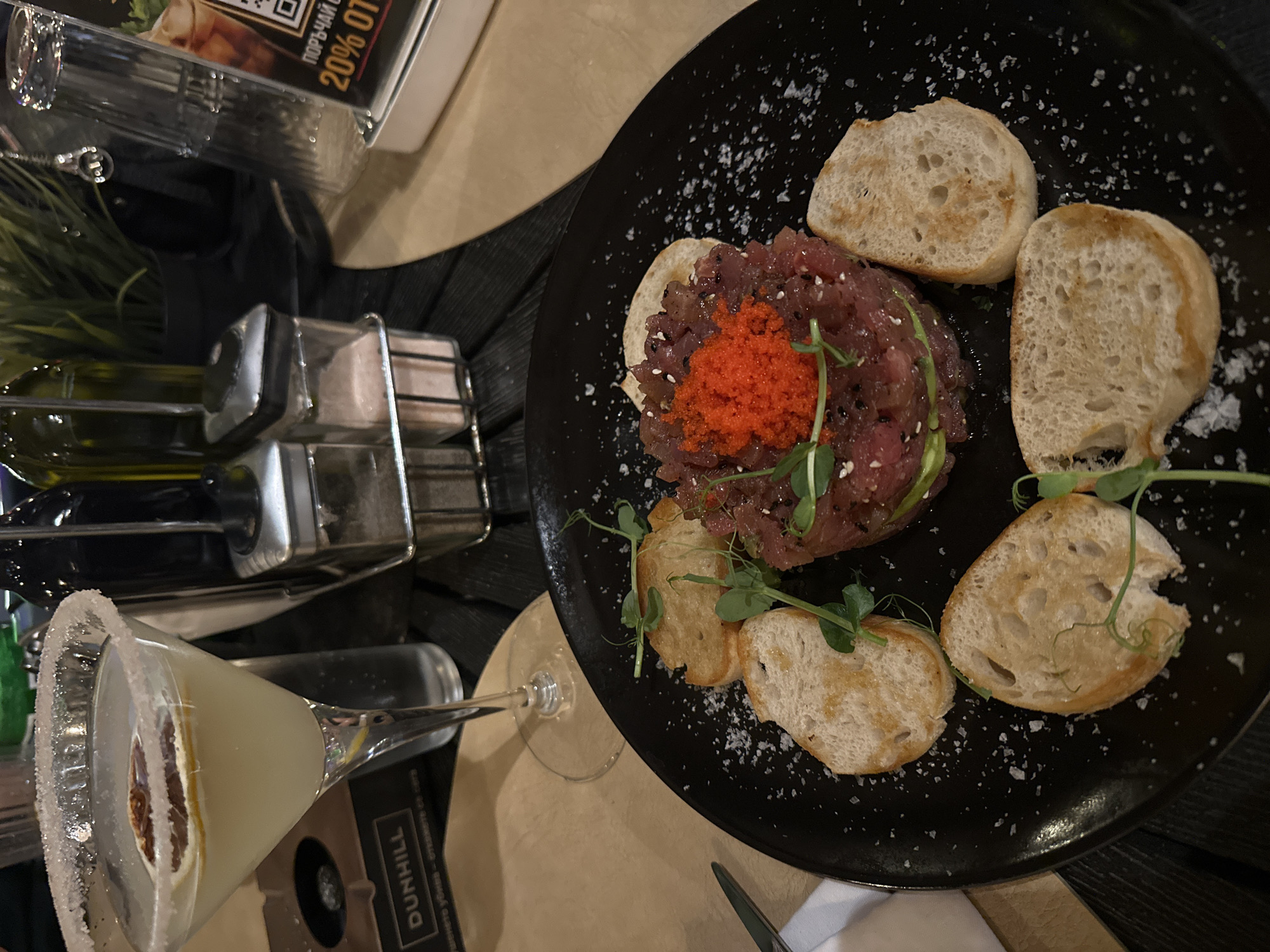 The image shows a close-up of a dish of tartare served with bread slices on a black plate, placed on a table with a dark tablecloth. A margarita cocktail with a salted rim and a lemon slice is visible in the background, along with salt and pepper shakers, and a glass. A menu or promotional flyer with Cyrillic text and the words "20% ОТ" (which translates to "20% OFF") is also present. The overall setting suggests a restaurant environment. There is no specific information in the image to indicate the location is Pazardzhik, Bulgaria.