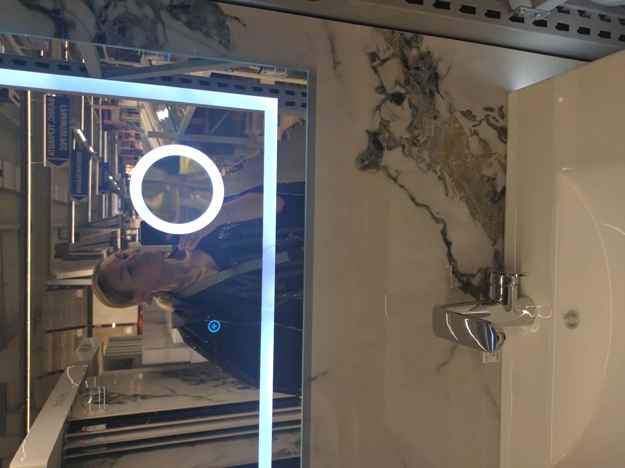 This image shows a bathroom sink and a marble countertop with a woman reflected in the mirror. The mirror has a lighted border and a circular light in the center. The faucet is chrome, and the sink is white. The marble wall has grey and gold veining. The reflection in the mirror shows a woman wearing a black top and a light-colored shirt underneath and what appears to be a store aisle.
Since this image was taken inside of a retailer store with display items for sale, it is impossible to pinpoint a specific place in the city of Stara Zagora, Bulgaria.
