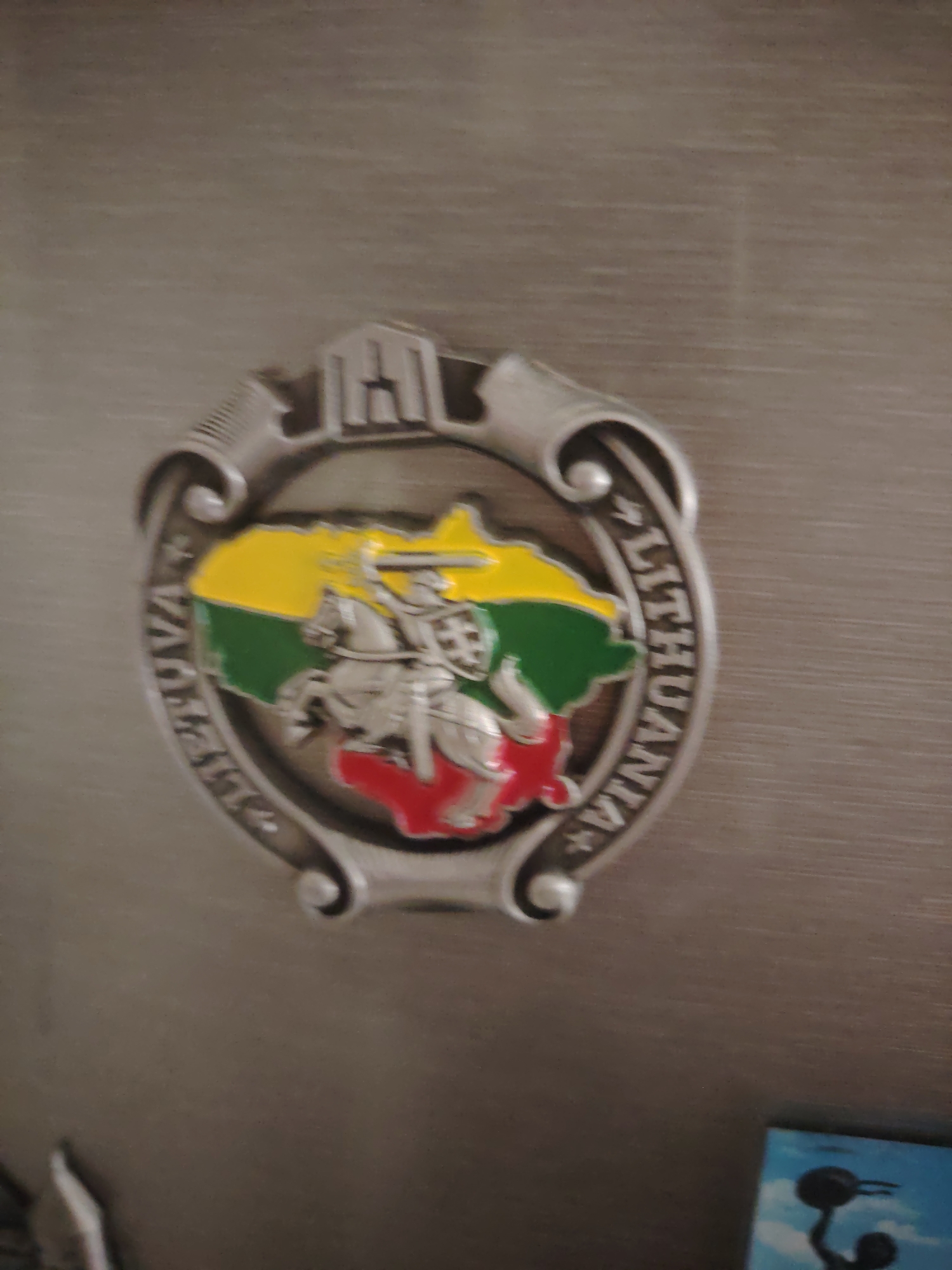 The image shows a close-up of a metallic emblem affixed to a textured, gray surface. The emblem is circular with decorative flourishes and features a map of Lithuania colored in yellow, green, and red. A knight on horseback, a depiction of the Vytis (the national coat of arms of Lithuania), is prominently displayed on the map. The word "LITHUANIA" is inscribed in capital letters around the outer edge of the emblem. The background is out of focus, but appears to be a brushed metal or laminate surface. There is a small, out-of-focus portion of a blue object with a silhouette of a person holding a weight visible in the bottom right corner of the image. There is no indication of the city of Huntingdonshire, United Kingdom, in the image.
