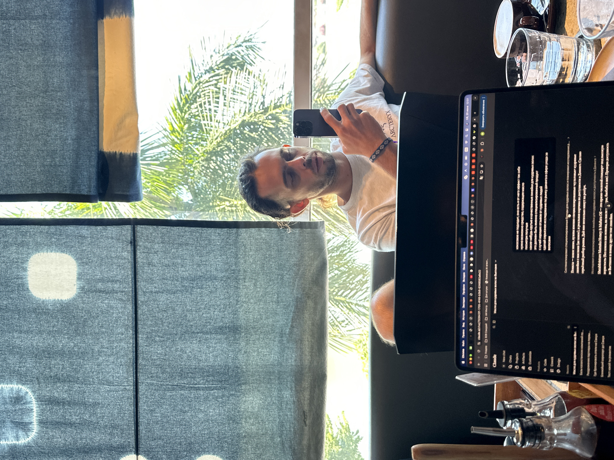 In the city of Đà Nẵng, Vietnam, a man with dark, curly hair pulled back sits in a dimly lit room, illuminated by natural light filtering through blinds patterned with white circles and a horizontal band. He is seated on a dark sofa and is holding a smartphone in his right hand, looking down at it. He wears a white t-shirt with the "ARC'TERYX" logo visible on the sleeve. In front of him, a laptop is open, displaying text and a user interface. The laptop screen shows a chat application with the name "Claude" and a list of recent activities. The text on the screen appears to be in Russian. To the right of the laptop, a glass of water and a dark mug are on a saucer. Behind the man, part of another person's head is visible. The environment suggests a cafe or a coworking space, with tropical foliage visible through the window, characteristic of Đà Nẵng. A wooden container with two bottles of condiments is visible in the foreground to the left.