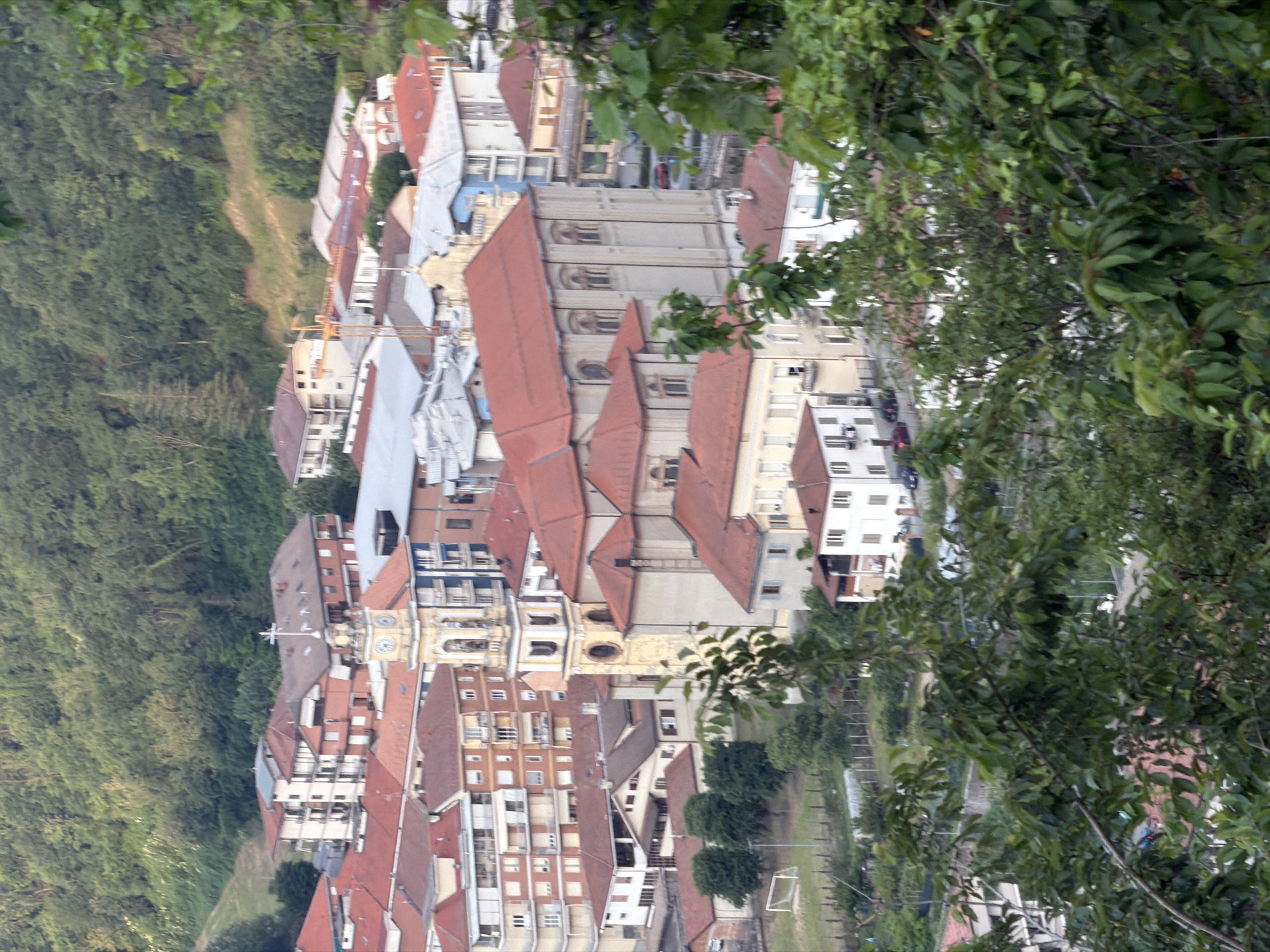 The image shows an aerial view of a European town. Predominantly, the town features buildings with red tile roofs and light-colored walls, likely made of stone or concrete. A large church dominates the center of the town, characterized by a basilica-style structure with a bell tower. The town is nestled among green trees, especially lush on the left side of the image, which suggest a forest or wooded area. The town appears to be in a valley or on a hillside. A soccer field is to the right of the church, further down the hill. The image is taken from an elevated position, possibly a higher hill or a nearby mountain. The foreground contains foliage, which provides a frame for the shot.