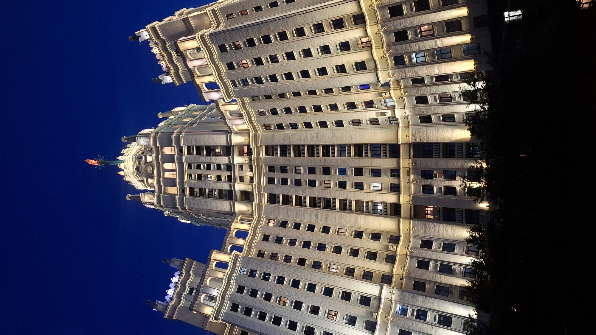 An upward shot captures a towering, multi-story building illuminated at night. The building is constructed of light-colored stone and features numerous windows, many of which are lit from within. The architecture is ornate, with decorative elements and a prominent spire at the top, topped with a red beacon. The sky is a deep, clear blue. The lower portion of the image is obscured by dark silhouettes of trees and other indistinct objects.