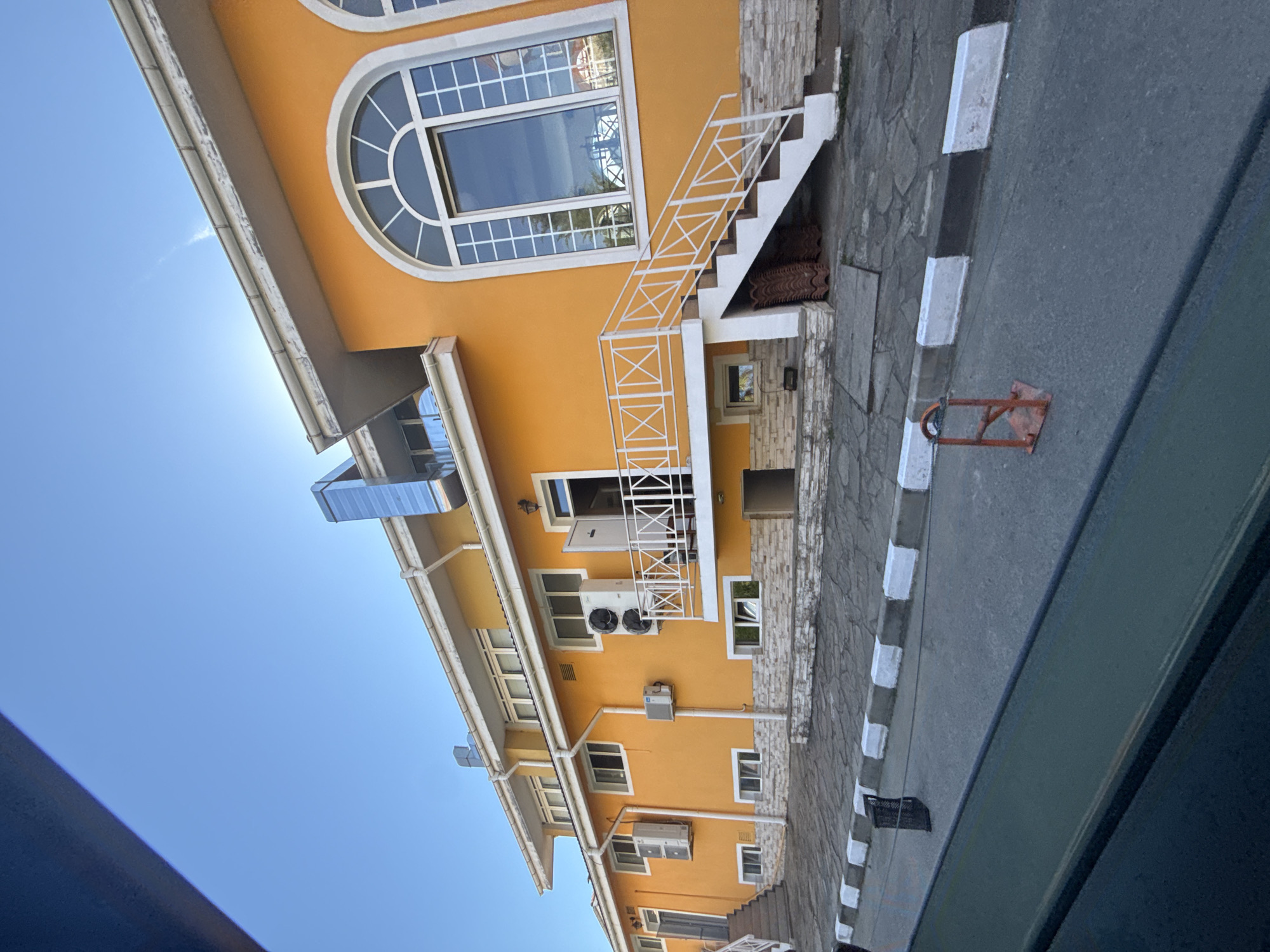 The image depicts a two-story, bright orange building with white trim and a white railing on a balcony. The building has several windows, including a large arched window on the second floor. There are two outdoor air conditioning units visible on the exterior walls, and a metal ventilation pipe extends upwards from the roof. A white staircase with a white railing leads to the balcony. The ground in front of the building is paved with stone, and a sidewalk with black and white painted curbs runs along the street. A red parking barrier is visible on the asphalt road. The sky is clear and blue. The building appears to be a commercial property or residence located in the city of Sozopol, Bulgaria.