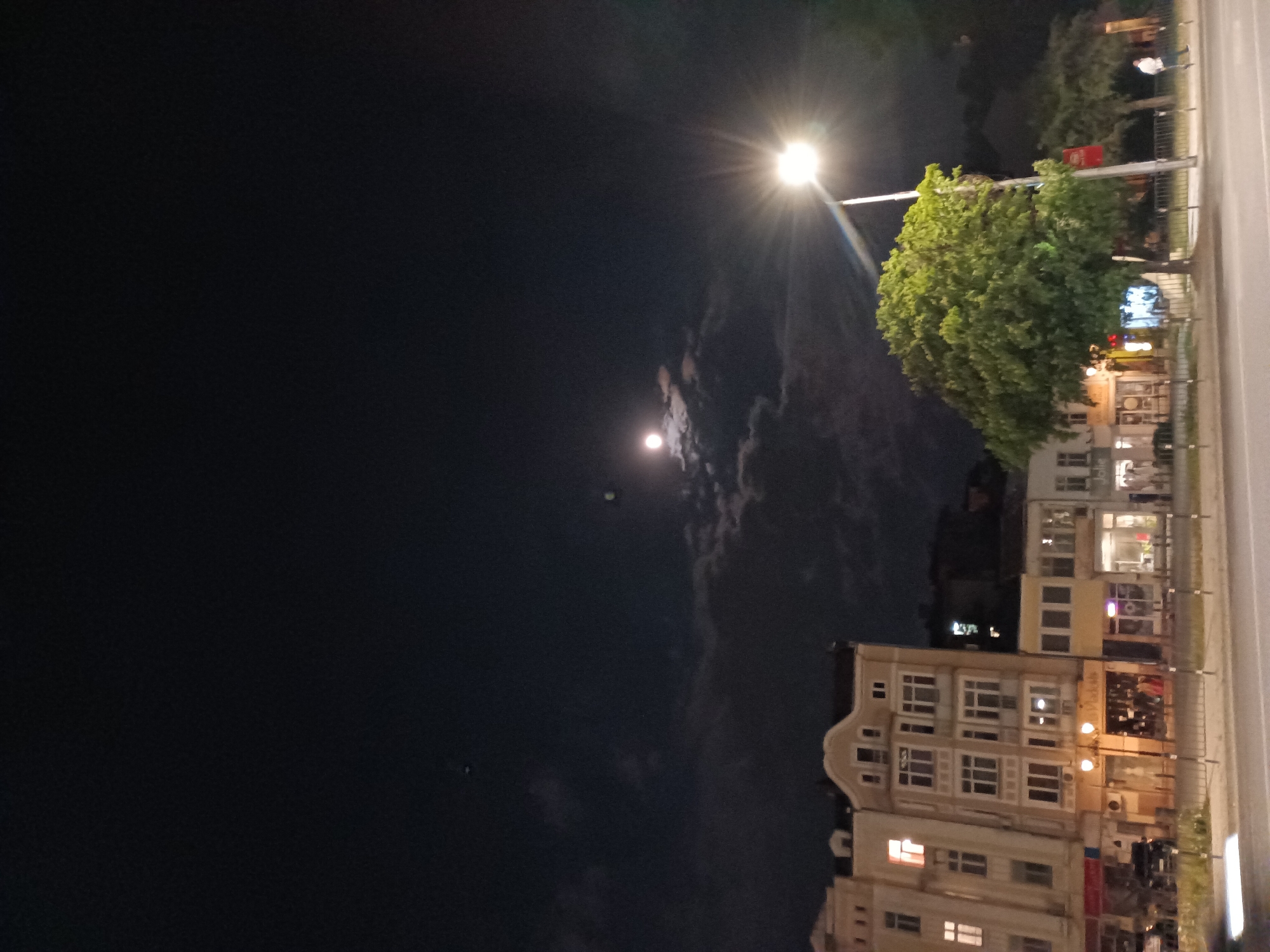 This is a nighttime, eye-level, vertical, outdoor shot of a street scene in Plovdiv, Bulgaria. The image is dominated by a dark sky with clouds, a bright street lamp, and a moon. The street lamp is in the upper right quadrant of the image, casting a bright light with visible rays. The moon is visible in the upper left quadrant, appearing as a bright orb partially obscured by clouds. Below the sky, a row of multi-story buildings lines the street. The buildings are illuminated, with some windows showing interior lights, suggesting it is evening or night. The architectural style of the buildings appears to be mixed, with some having more modern facades and others exhibiting older features. Green trees are also present, with one large, lush tree prominently displayed in the middle right of the frame. A street or sidewalk runs alongside the buildings, bordered by a metal fence. There is a hint of traffic or parked vehicles visible in the lower right portion of the image. The overall mood is calm and urban, illuminated by artificial and natural light sources in the dark. There is some visible text on signage on the buildings, but it is not clearly legible.